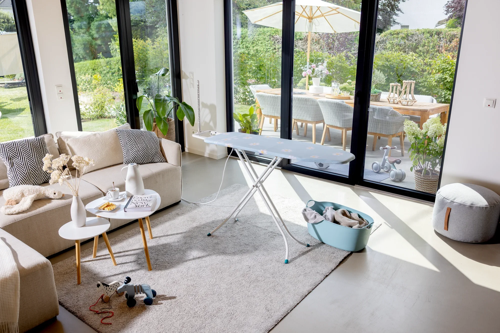 Bügelbrett Leifheit Classic Basic Plus in einem modernen Wohnzimmer mit großen Fenstern. Es steht auf einem Teppich neben einem Tisch mit Dekorationen und einem Spielzeug.