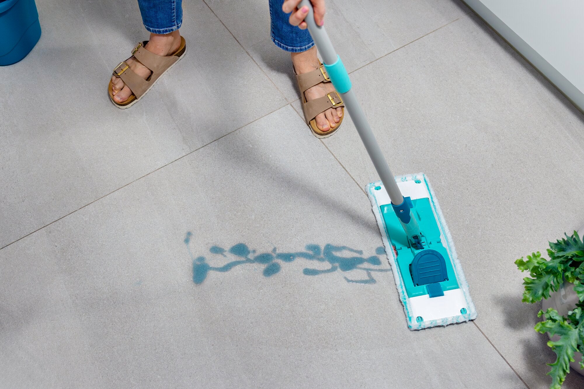 A person cleans a light grey tile floor with the POWER CLEAN system. The mop has a turquoise design and effectively removes dirt and stains. Plants can be seen in the foreground, giving the room a fresh atmosphere.
