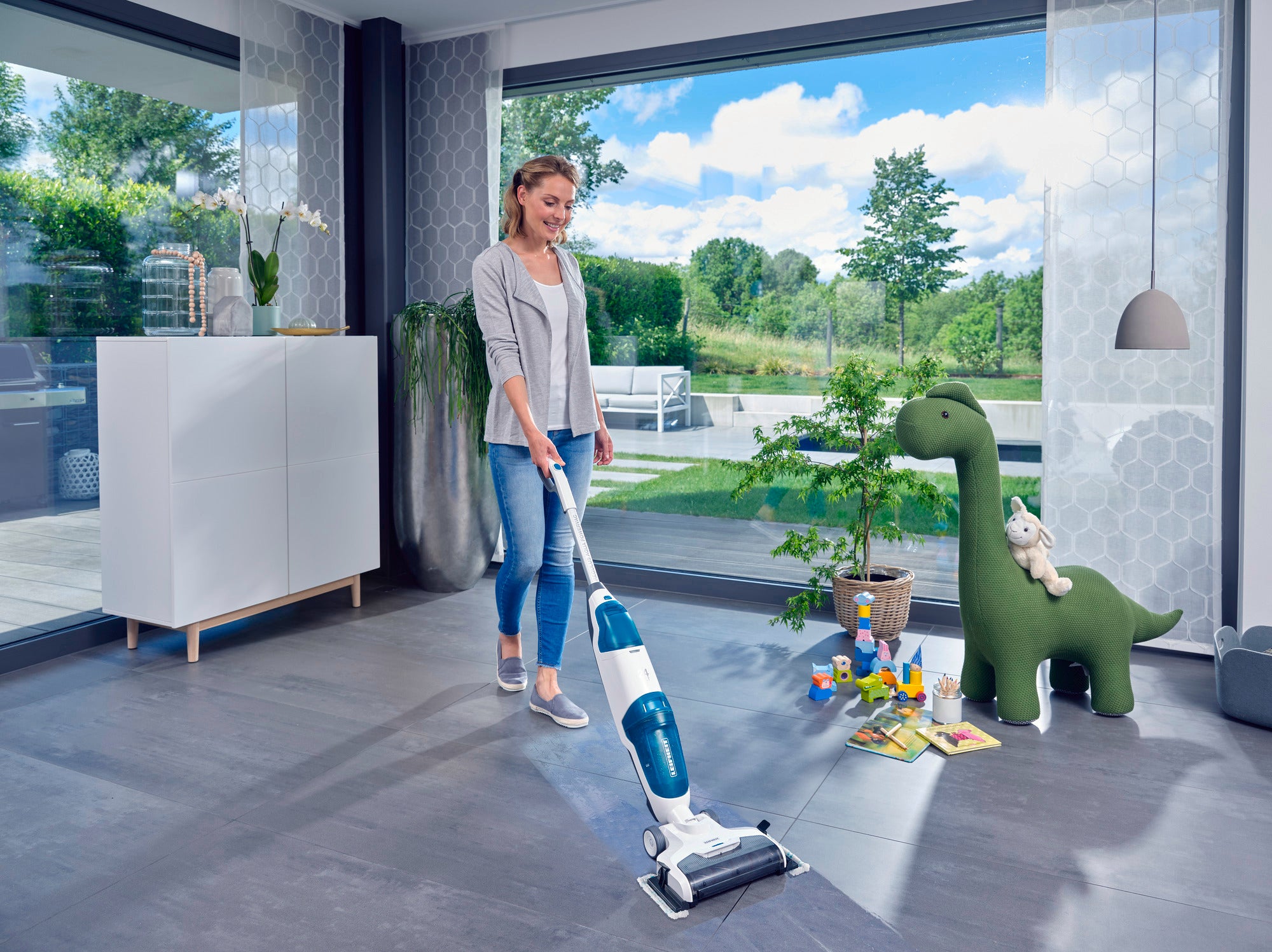 Eine Frau benutzt den Regulus Aqua PowerVac Pro, um den Boden in einem modernen Wohnzimmer zu reinigen. Im Hintergrund sind große Fenster mit Blick auf einen Garten.