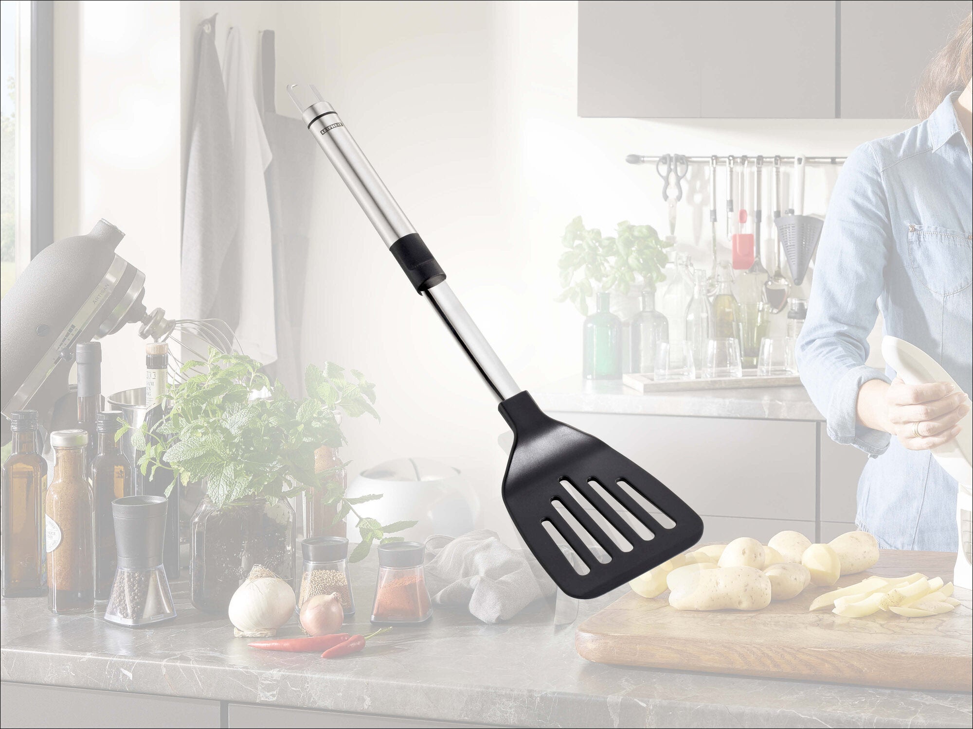 Leifheit Küchenhelfer Pfannenwender ProLine mit Edelstahlgriff und schwarzem, geschlitztem Wenderkopf, freigestellt vor heller Küchenszene mit Kochperson, Schneidebrett und frisch geschnittenen Kartoffeln.