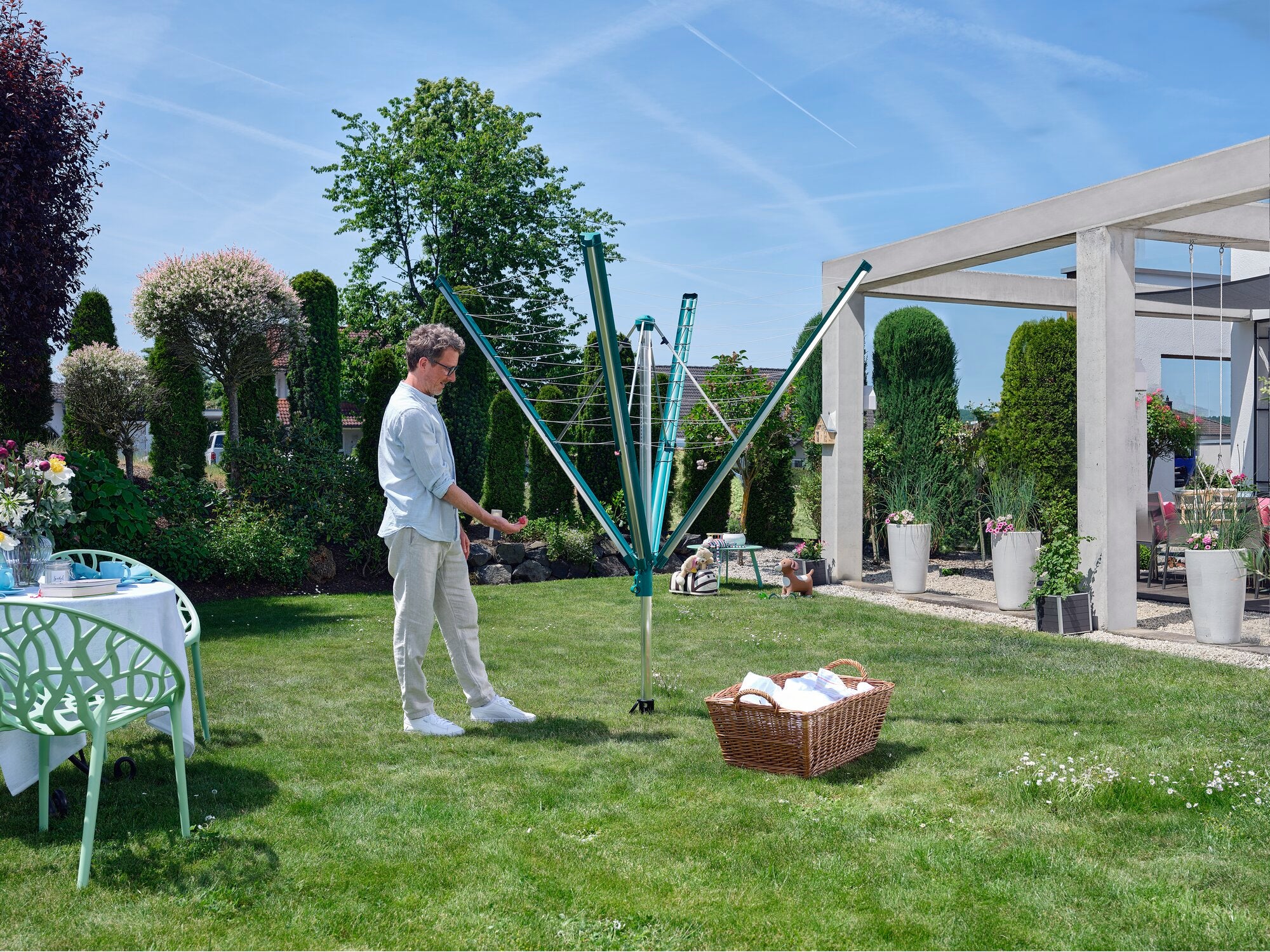 A man stands in a well-kept garden using the Linomatic 500 Deluxe rotary clothes dryer. The rotary clothes dryer is open and ready for hanging laundry. Plants and a modern gazebo can be seen in the background.