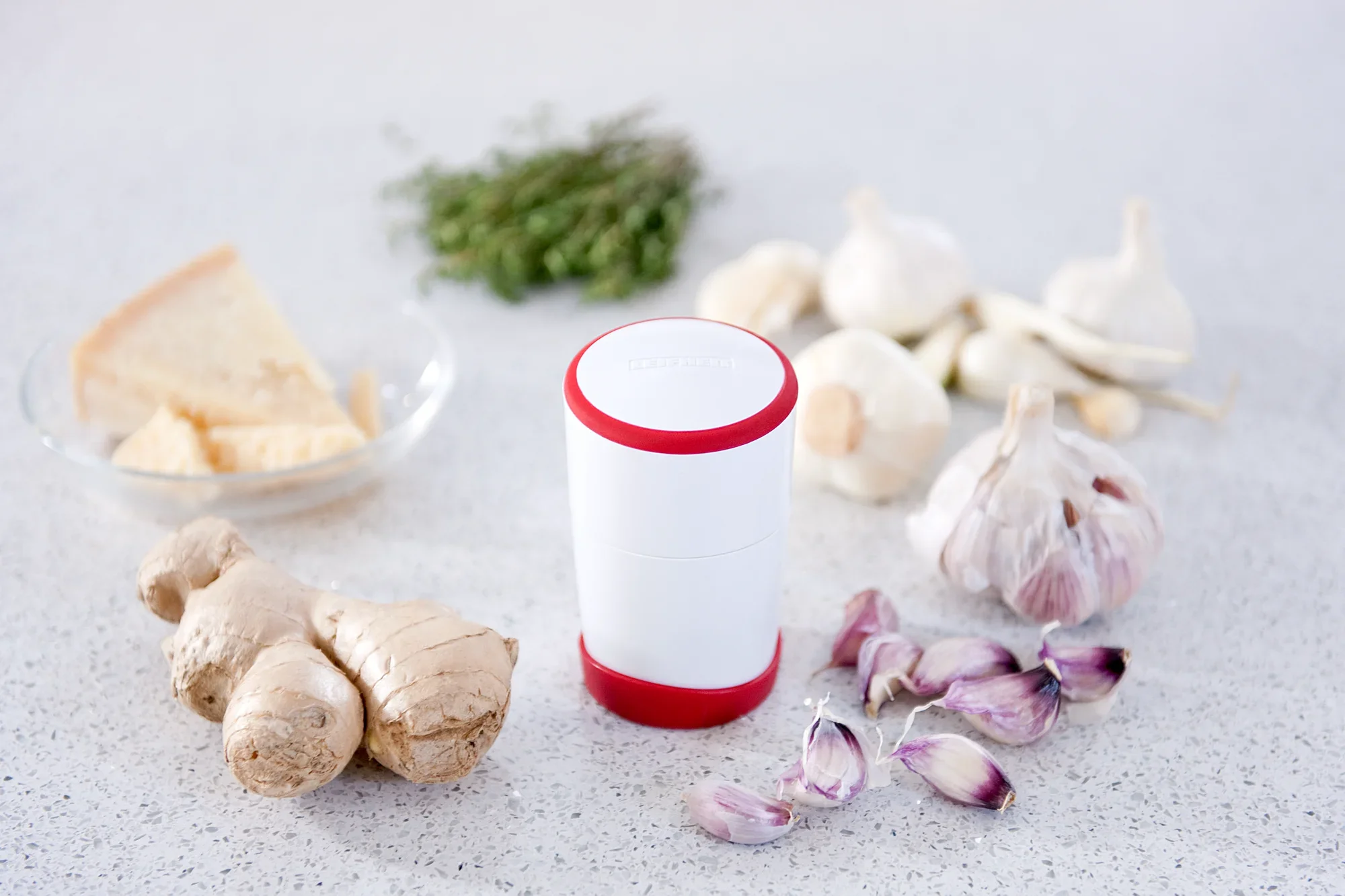 Een knoflooksnijder met een rode rand staat op een keukenblad. Er omheen liggen verse knoflookteentjes, gember, kaas en kruiden, die een uitnodigende kooksfeer creëren.
