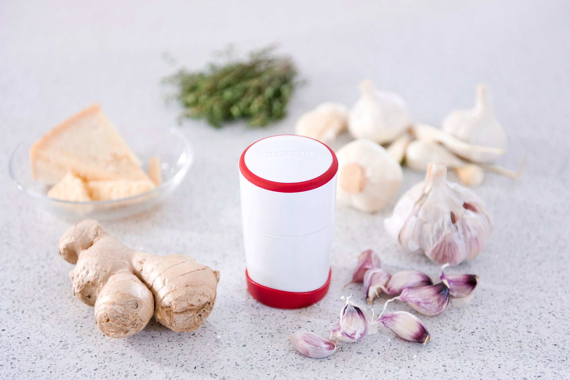 Un coupe-ail avec un bord rouge est posé sur un plan de travail. Autour de lui se trouvent des gousses d'ail frais, du gingembre, du fromage et des herbes, créant une atmosphère culinaire accueillante.