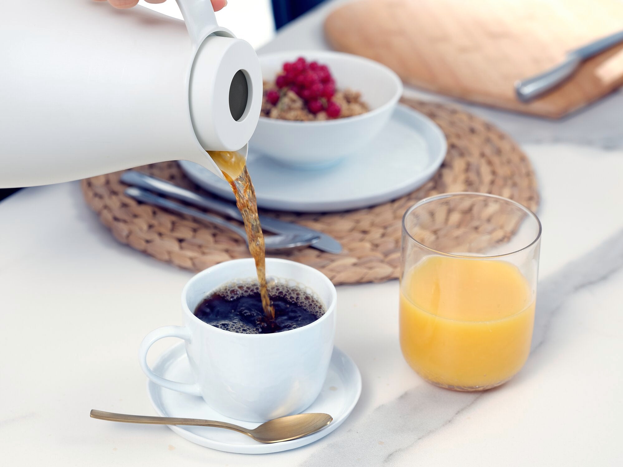 Isolierkanne Harmonic wird verwendet, um heißen Kaffee in eine weiße Tasse zu gießen. Im Hintergrund sind ein Glas Orangensaft und eine Schale mit Müsli zu sehen.