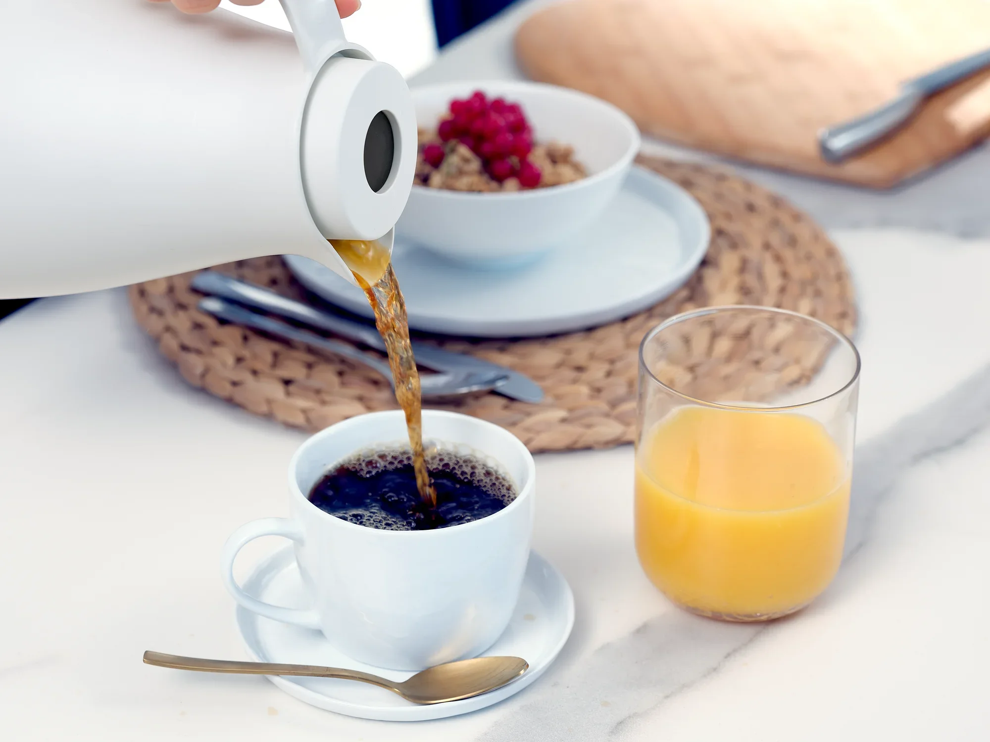 De Harmonic thermoskan wordt gebruikt om hete koffie in een wit kopje te schenken. Op de achtergrond staan een glas sinaasappelsap en een kom muesli.