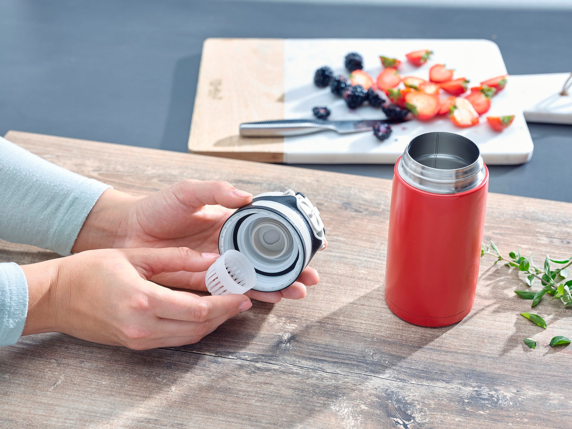 Isolierbecher Flip 350ml in Rot auf einem Tisch. Eine Person zeigt den abnehmbaren Deckel des Bechers. Im Hintergrund sind geschnittene Erdbeeren und Beeren zu sehen.