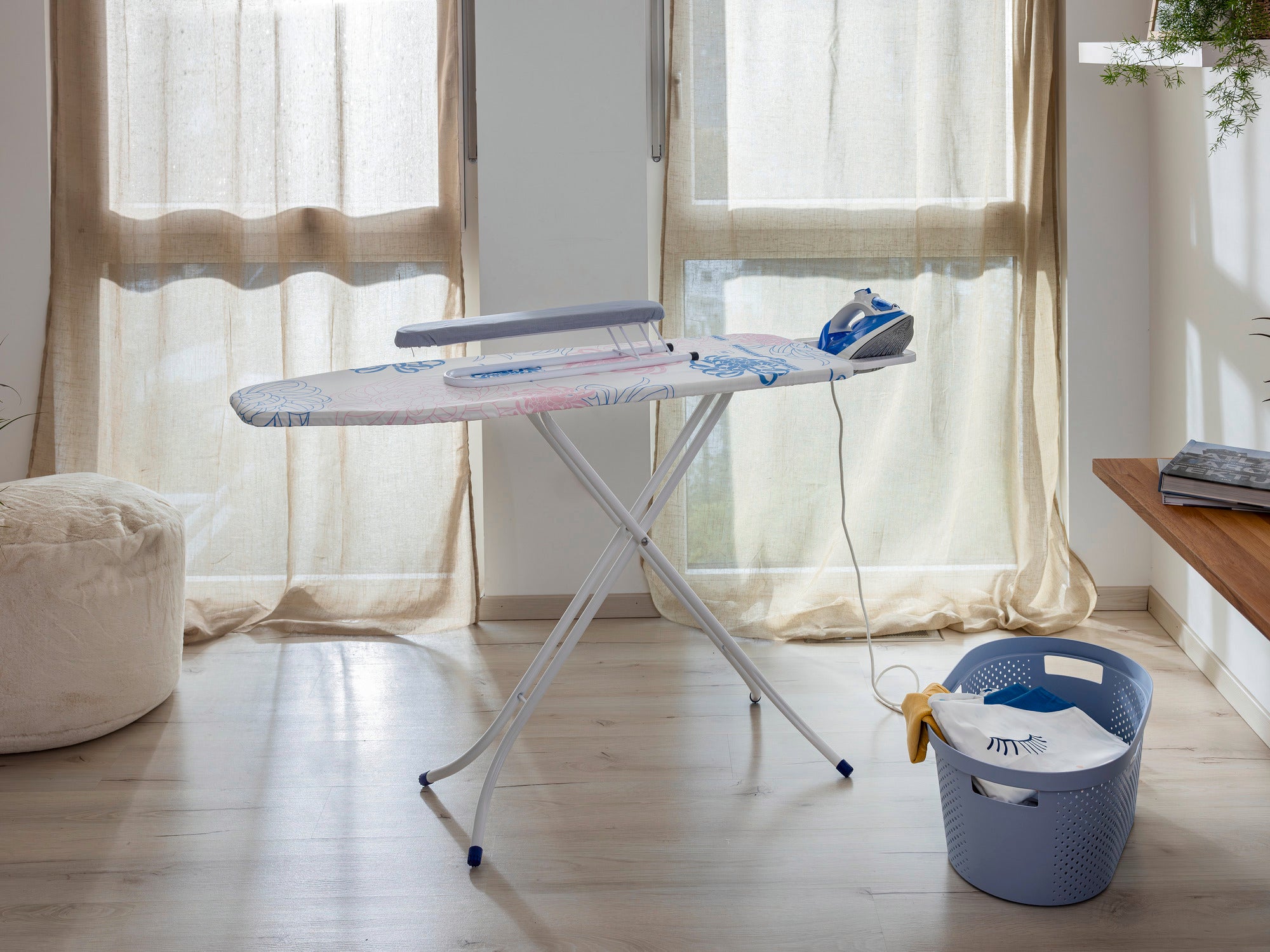 Una tabla para planchar mangas sobre una tabla de planchar en una habitación luminosa con grandes ventanas. Junto a ella hay una plancha y un cesto para la ropa. Ideal para planchar mangas con precisión.