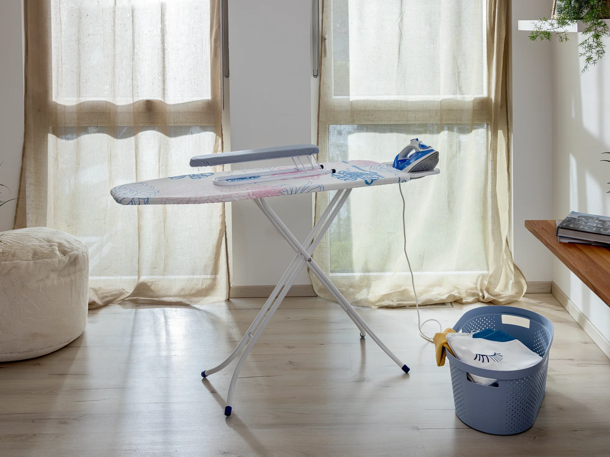 Een mouwplank op een strijkplank in een lichte kamer met grote ramen. Ernaast staat een strijkijzer en een wasmand. Ideaal om met precisie mouwen te strijken.