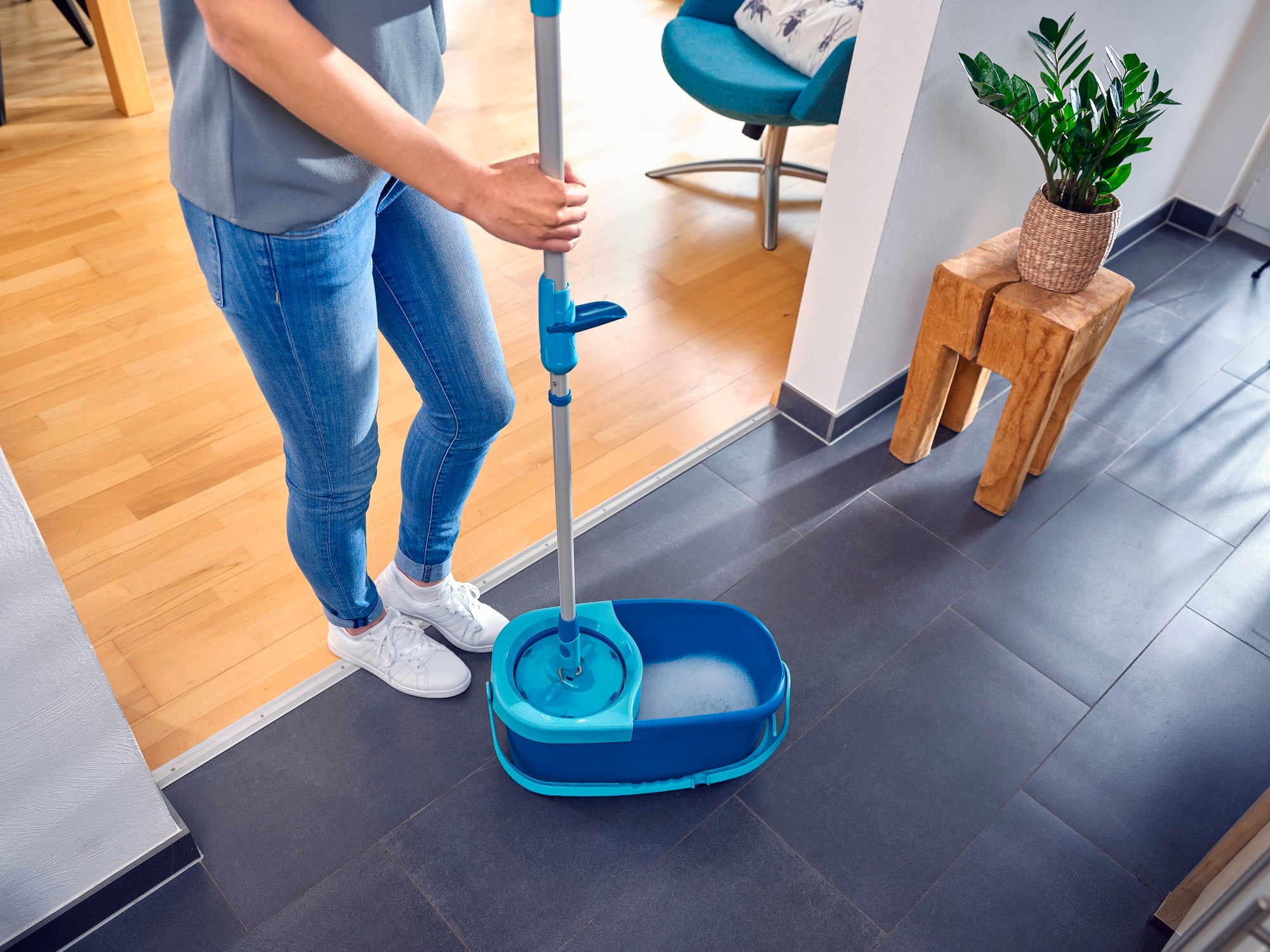 Eine Person reinigt einen dunklen Fliesenboden mit dem CLEAN TWIST Mop Ergo. Der Mop hat einen blauen Eimer und wird in einem modernen, hellen Raum verwendet. Ideal für effiziente Bodenreinigung.
