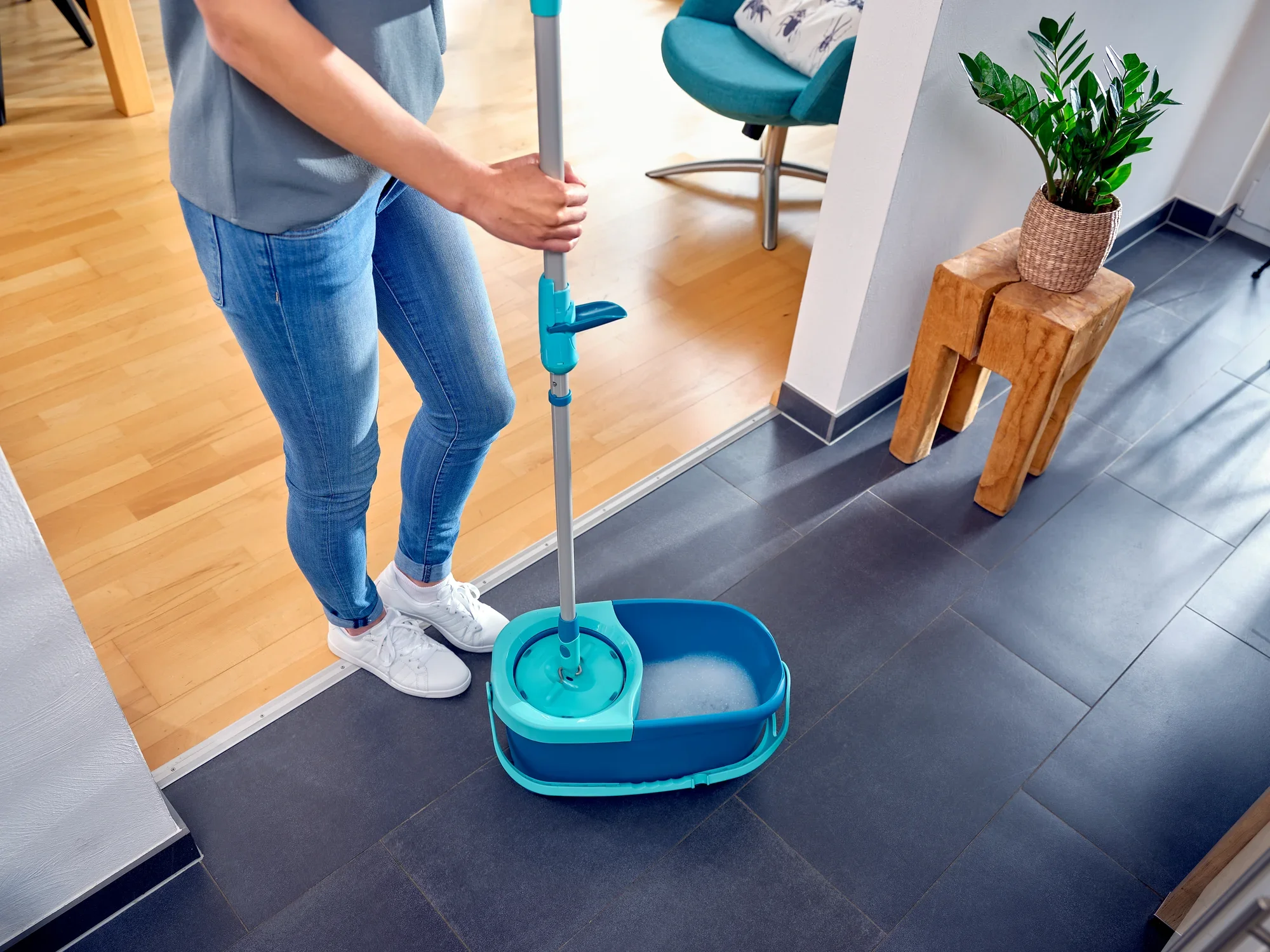 Una persona limpia un suelo de baldosas oscuras con la mopa CLEAN TWIST Ergo. La mopa tiene un cubo azul y se utiliza en una habitación moderna y luminosa. Ideal para una limpieza eficaz del suelo.