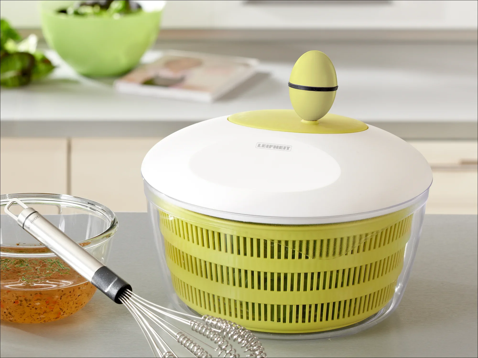 Wirówka do sałatek z zielonym sitkiem i białą pokrywką na blacie kuchennym. W tle widać miskę, trzepaczkę i książkę kucharską. Idealny do świeżych sałatek.