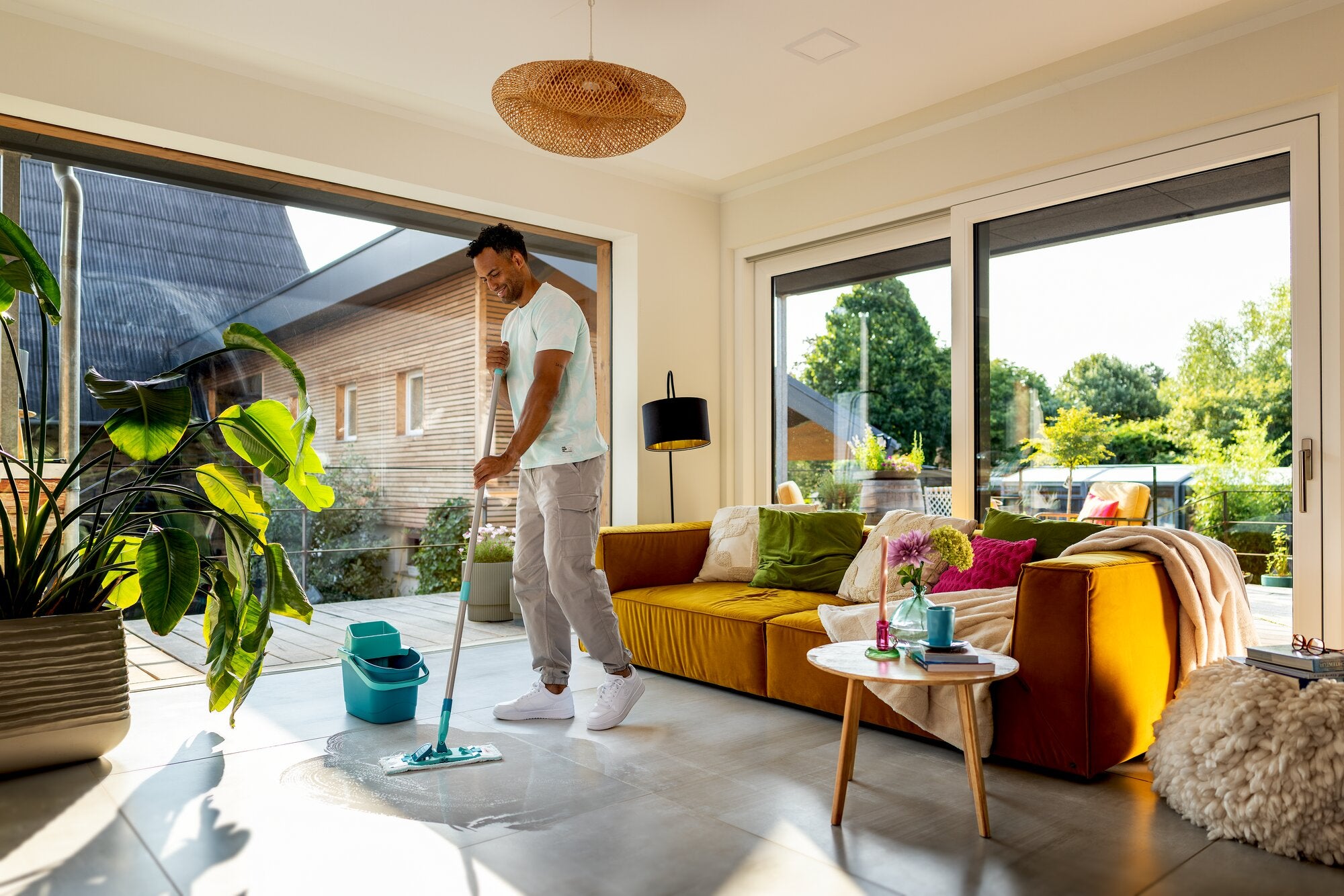 Leifheit POWER CLEAN lavapavimenti in Türkis im Einsatz: Ein Mann wischt helle Fliesen in einem sonnigen Wohnzimmer mit großem Fenster, Sofa, Pflanzen und modernem Wohnambiente.