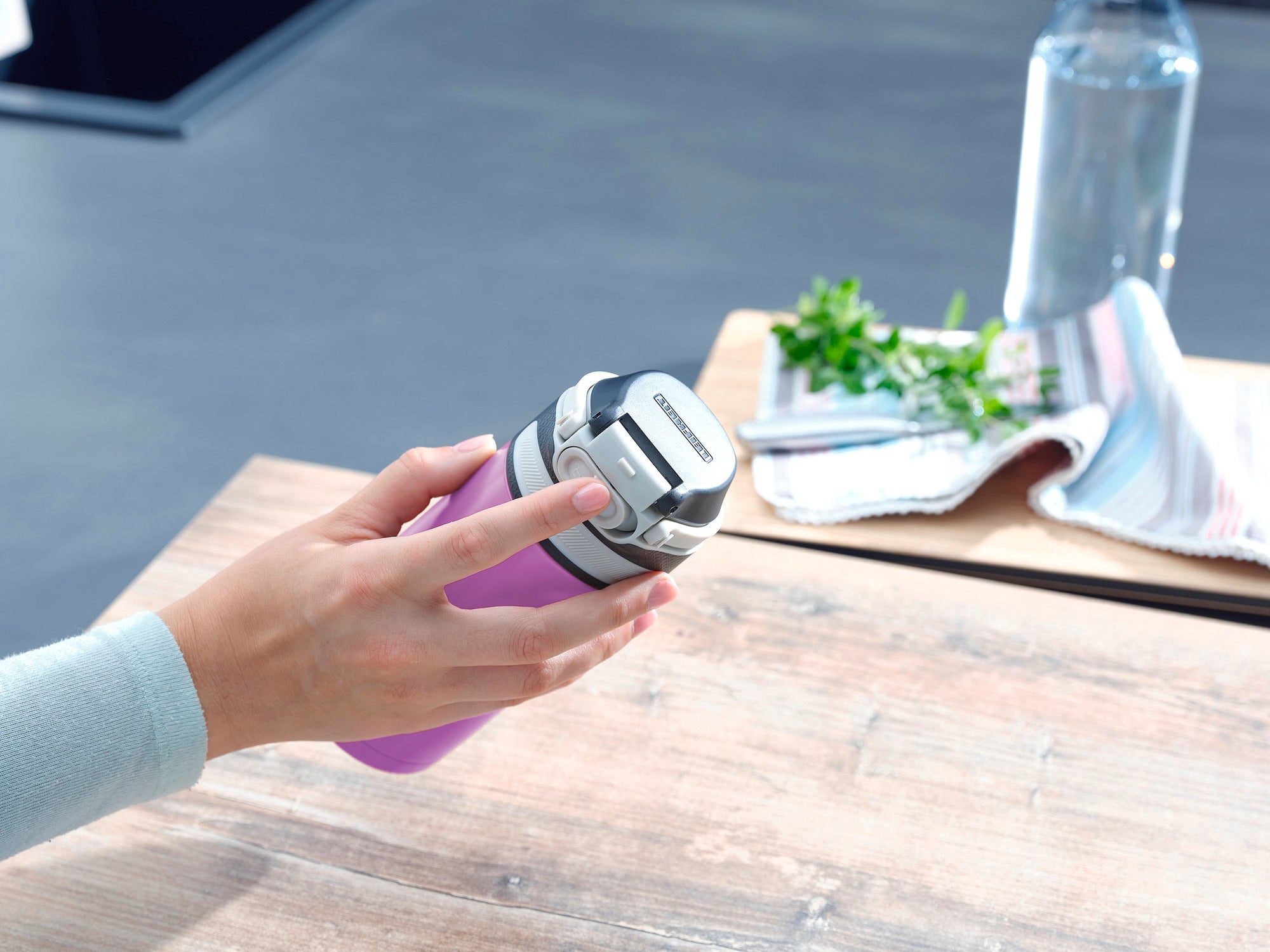 Una persona tiene in mano un thermos Flip viola da 350 ml. Il thermos ha un coperchio argentato ed è appoggiato su un tavolo di legno accanto a un asciugamano e una bottiglia d'acqua.