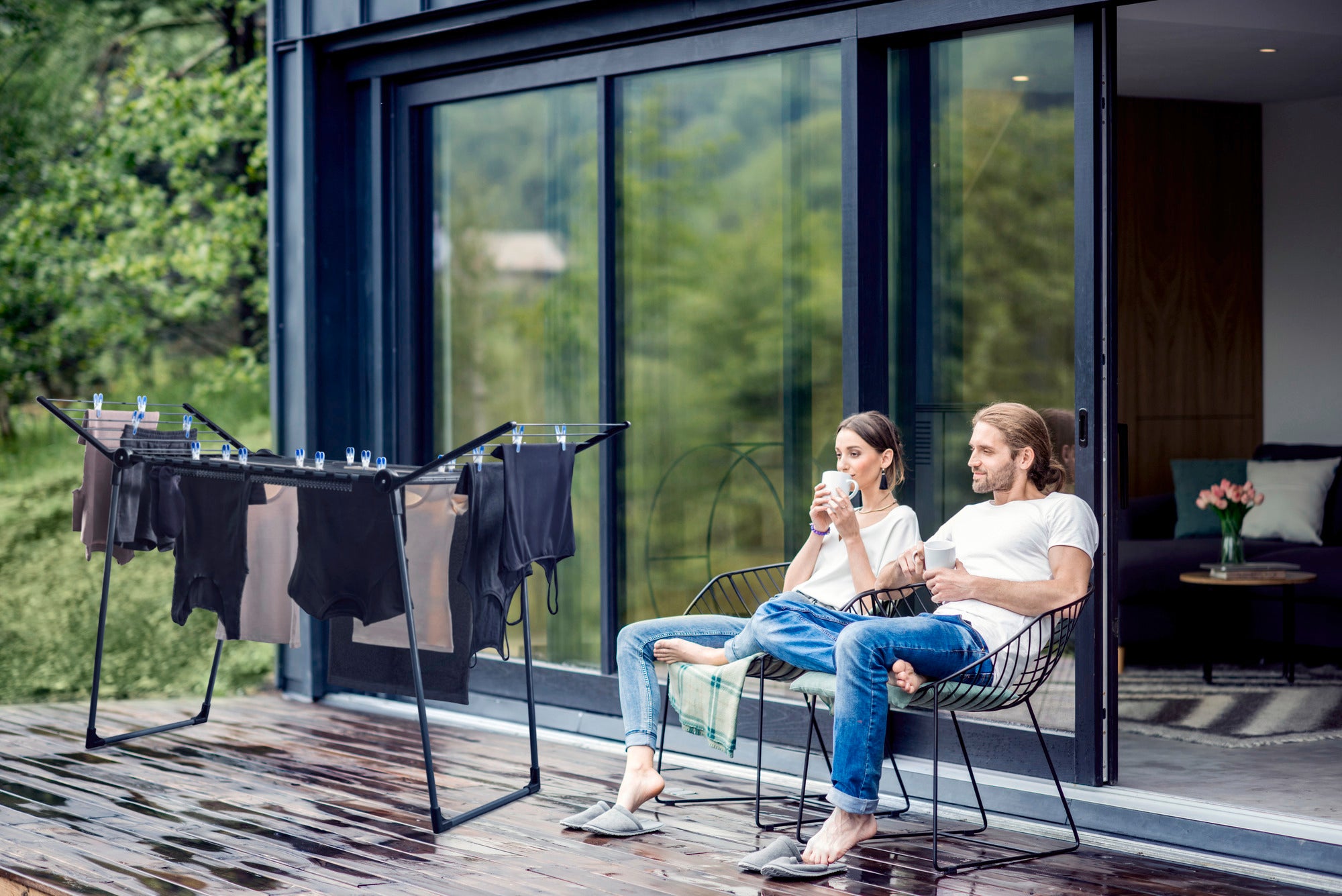 Un couple est assis, détendu, sur une terrasse devant une maison moderne. À côté d'eux se trouve le sèche-linge sur pied Pegasus 180 Solid, chargé de vêtements fraîchement lavés.