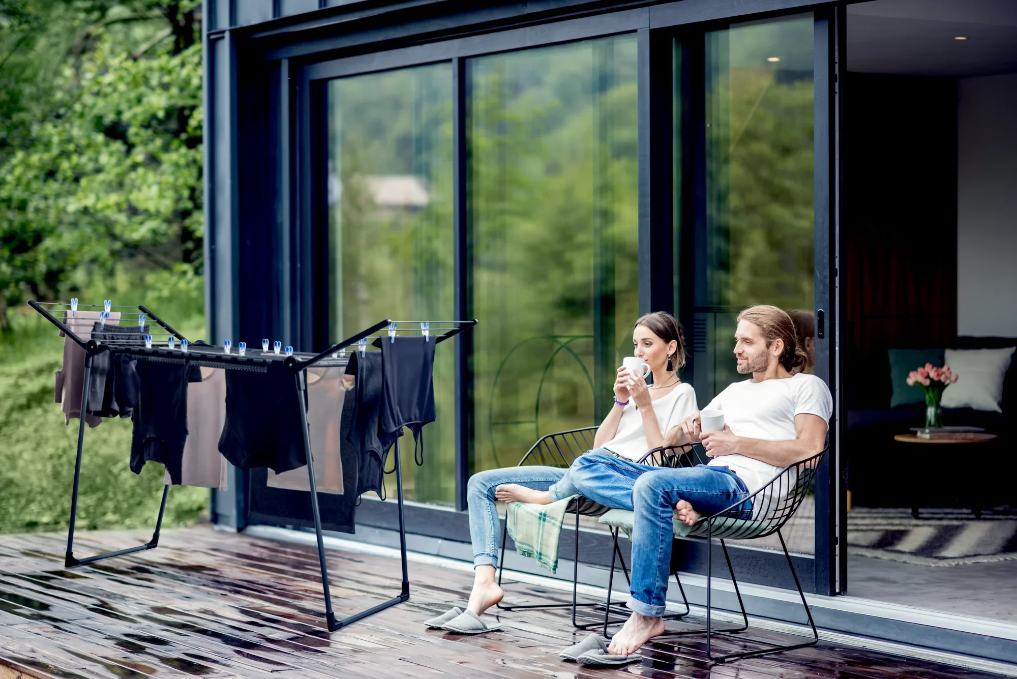 Para siedzi relaksując się na tarasie przed nowoczesnym domem. Obok nich znajduje się wolnostojąca suszarka bębnowa Pegasus 180 Solid, załadowana świeżo wypranymi ubraniami.