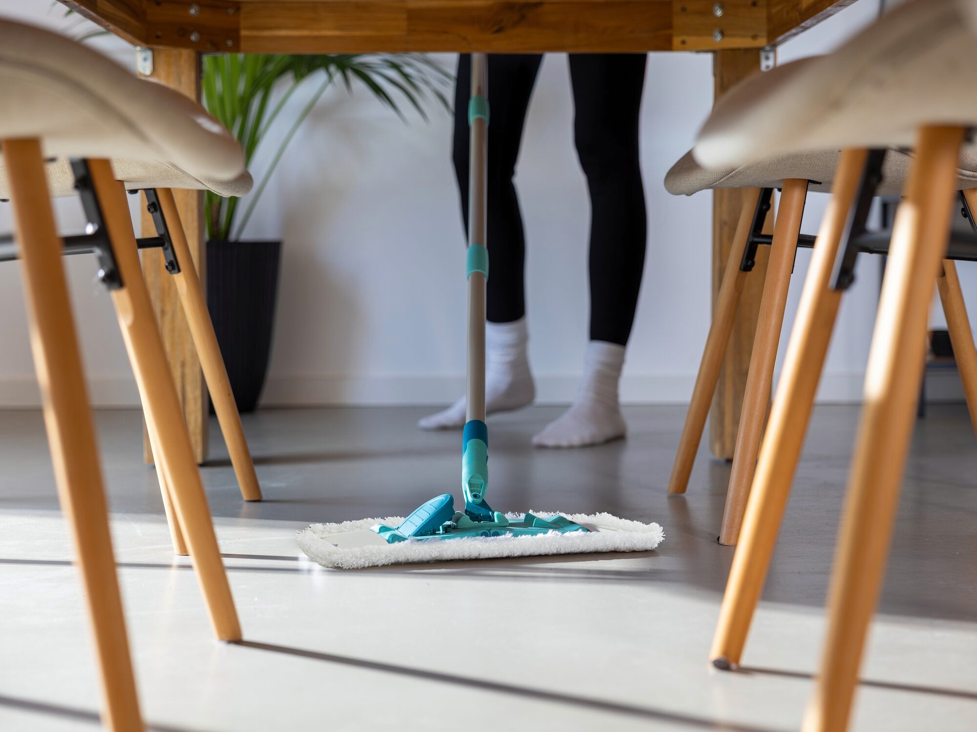 Un balai à franges modèle SUPERDUSTER est utilisé sous une table. Une personne nettoie le sol avec le balai à franges, tandis que des plantes sont visibles en arrière-plan.