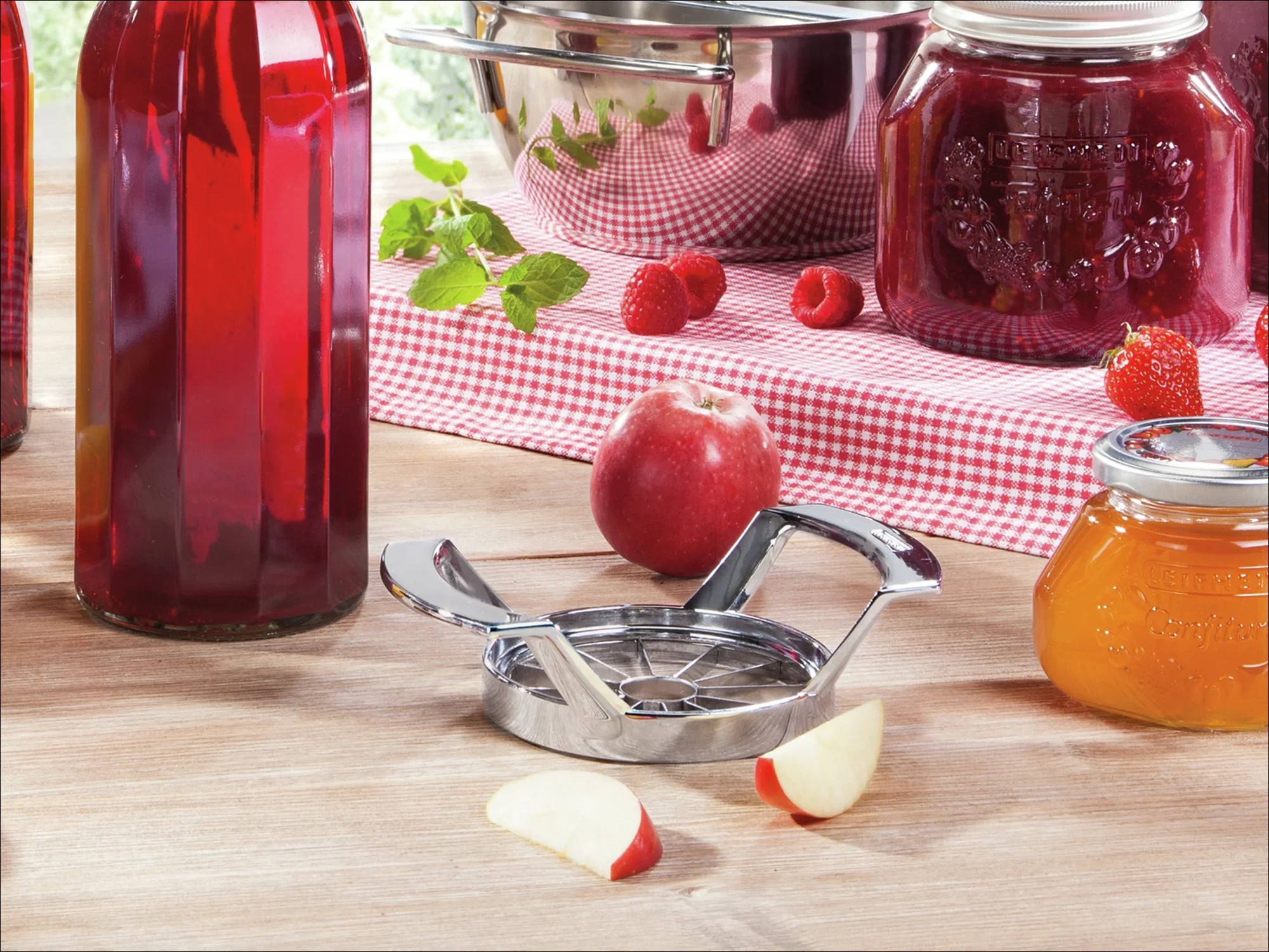 Krajalnica do jabłek ze stali nierdzewnej leży na drewnianym stole w otoczeniu czerwonych butelek, jabłka, słoików z dżemem i świeżych malin na czerwono-białej kraciastej tkaninie.
