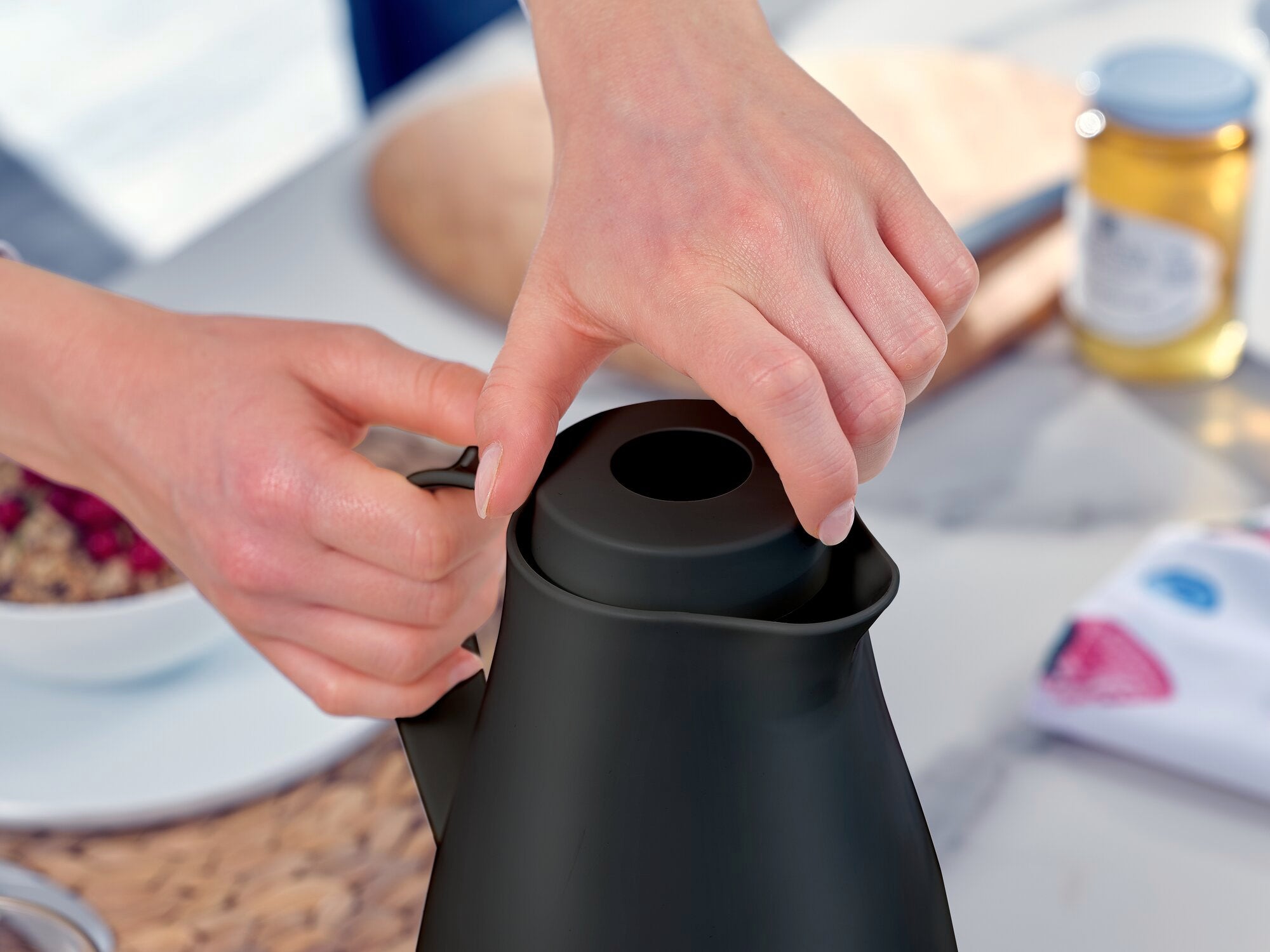 Een persoon opent het deksel van de zwarte "Harmonic" isoleerkan. Een glas honing en een kom muesli zijn onscherp te zien op de achtergrond. De scène straalt een gezellig ontbijtmoment uit.