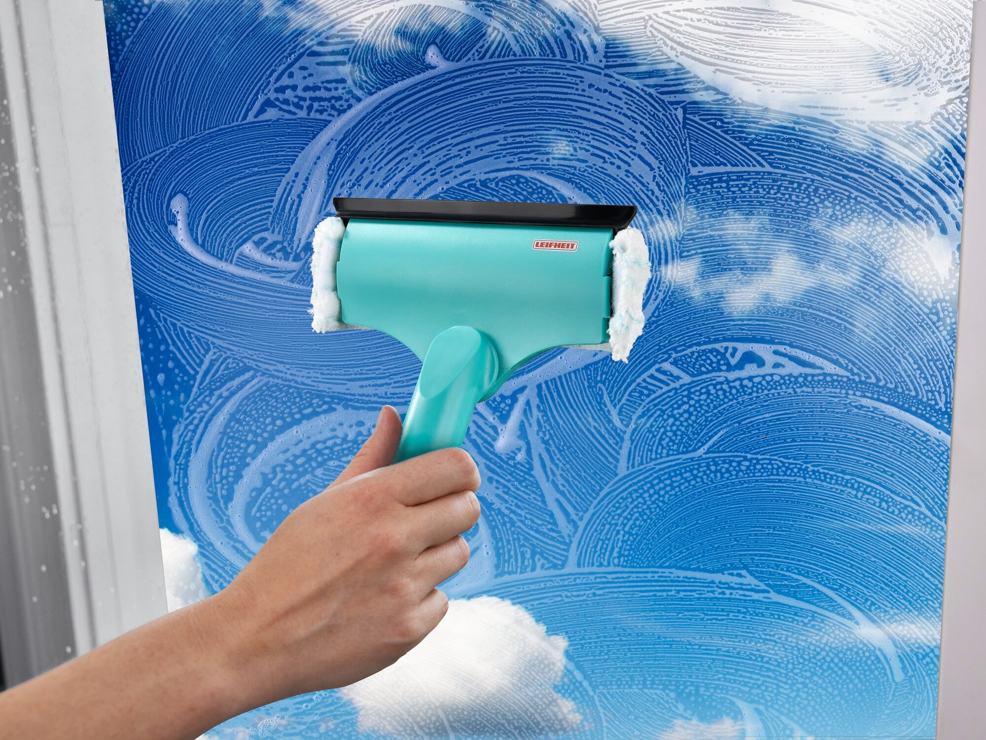 A 3-in-1 mini window and frame wiper is used to clean a window. The wiper has a turquoise handle and a black rubber lip.