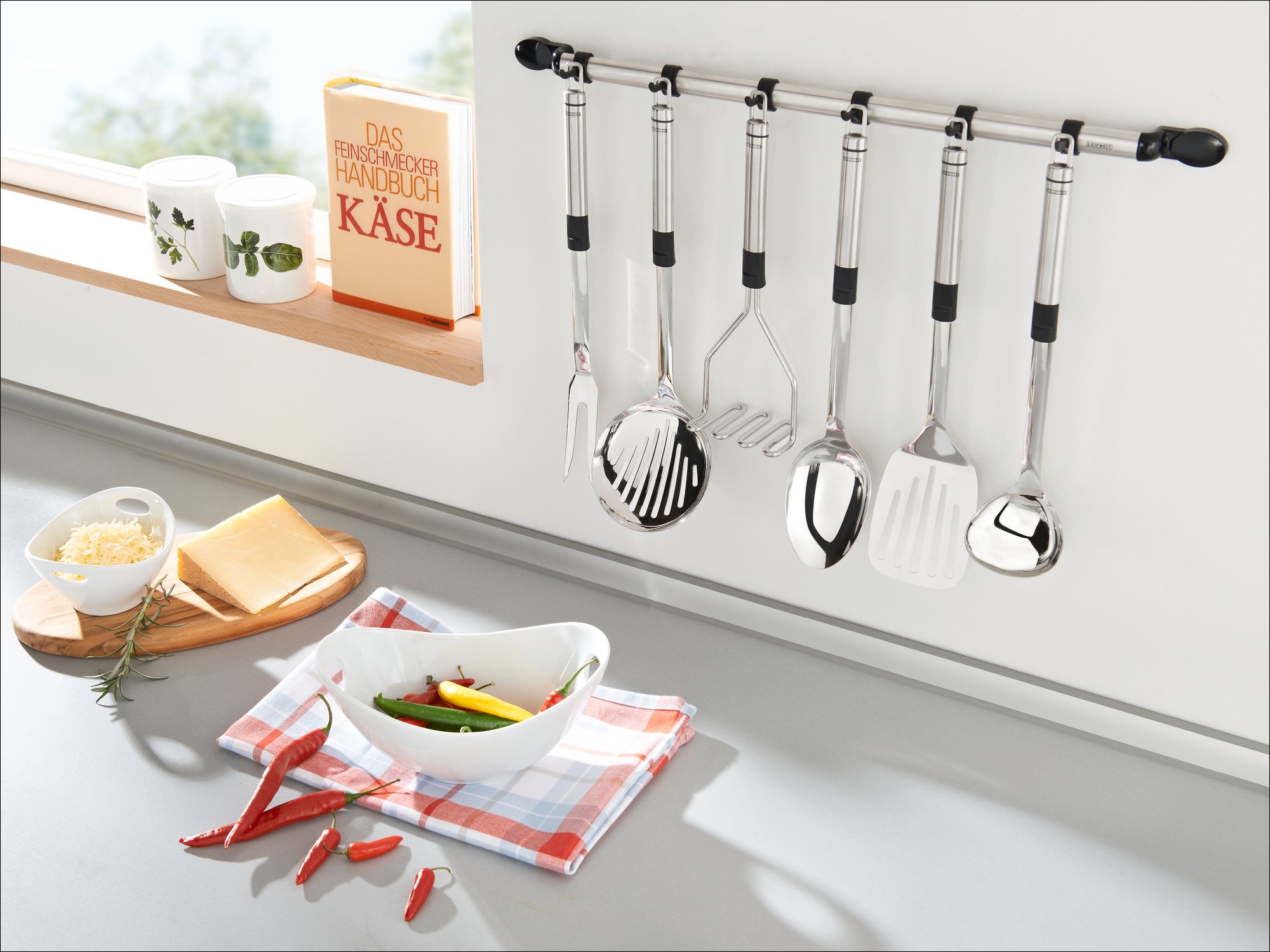 Eine moderne Küche mit einer Reihe von Edelstahl-Küchenutensilien, die an einer Wandhalterung hängen. Darunter befinden sich ein Schneidebrett mit Käse und Paprika sowie eine Schüssel mit Gemüse.