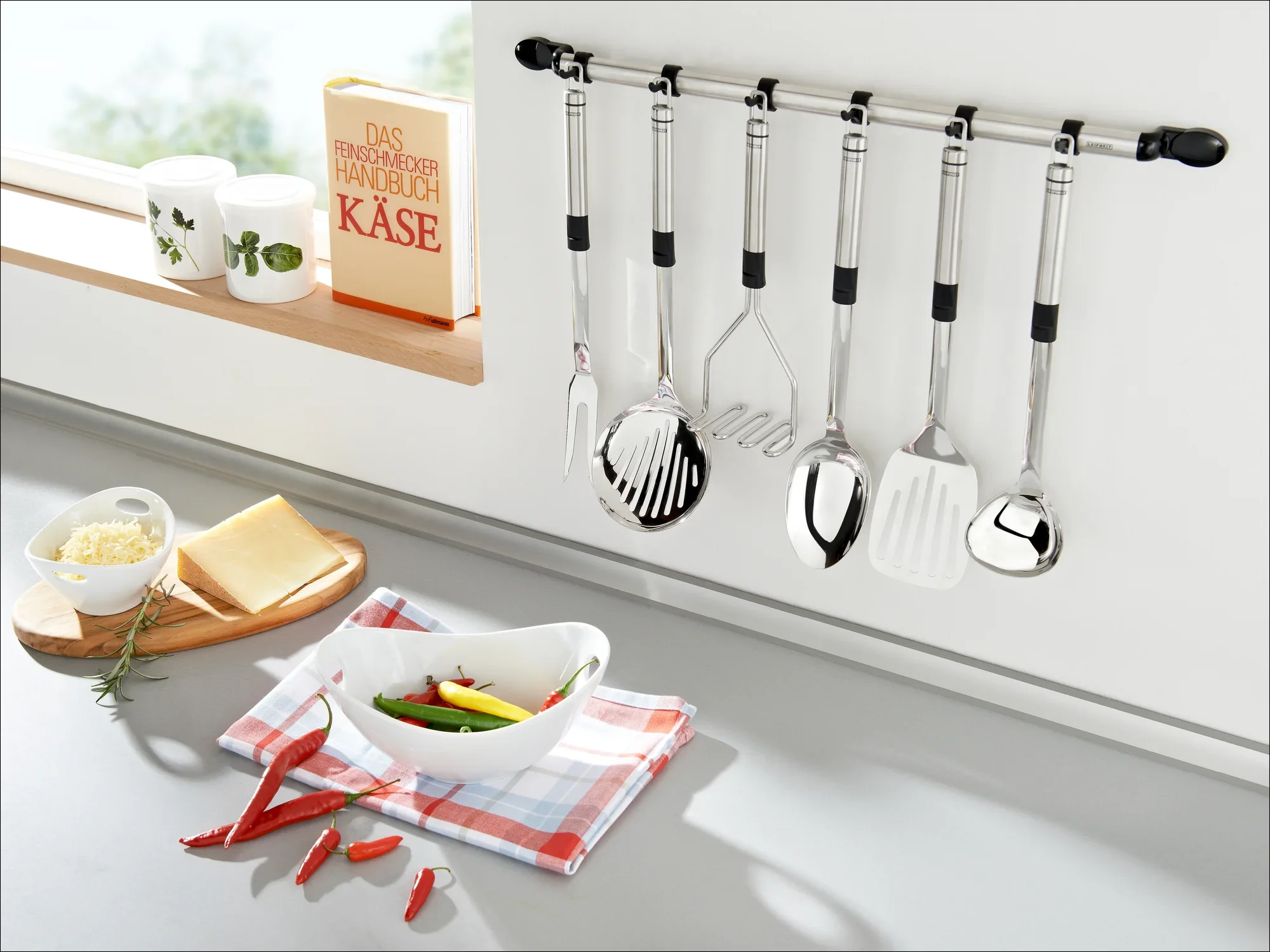 Een moderne keuken met een reeks roestvrijstalen keukengerei aan een muurbeugel. Dit zijn onder andere een snijplank met kaas en paprika en een schaal met groenten.