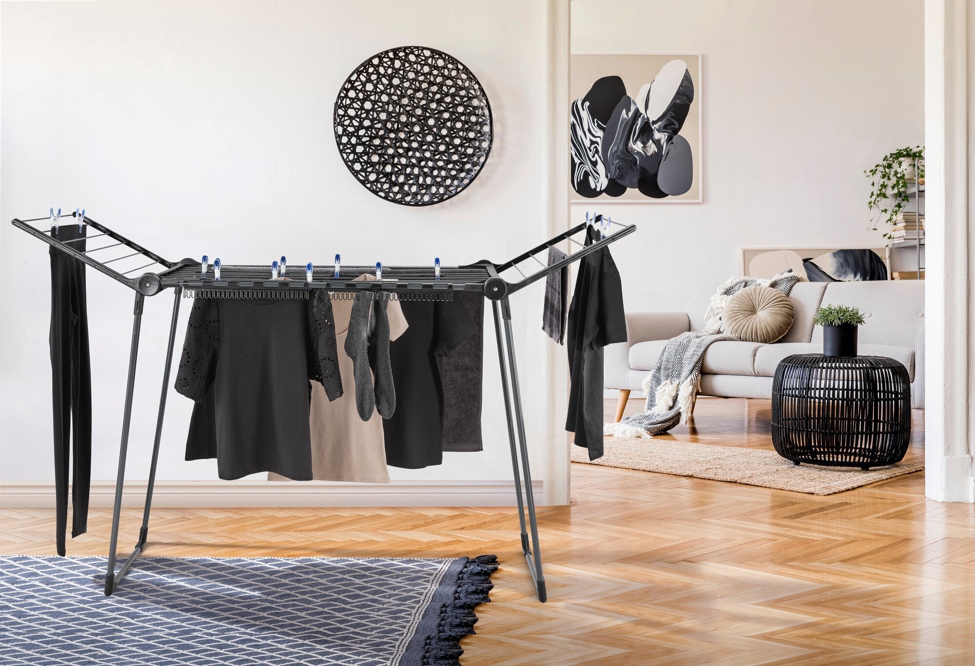 Ein Standtrockner Pegasus 150 Solid Slim Black steht in einem modernen Wohnzimmer. Der Trockner ist mit verschiedenen Kleidungsstücken behängt und passt perfekt in die stilvolle Umgebung.