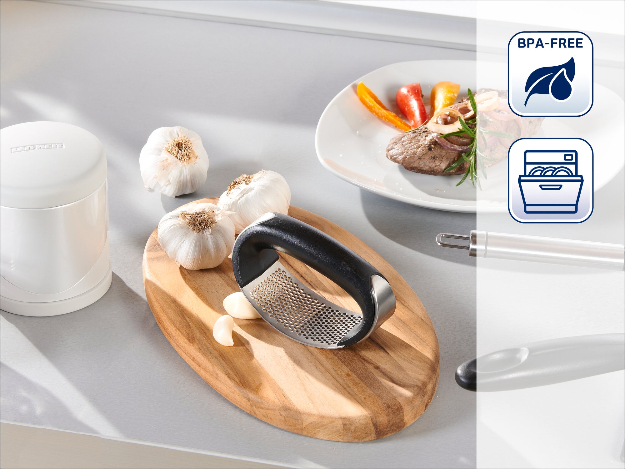 Garlic press on a wooden board with fresh garlic. In the background, a plate with meat and vegetables. Symbols indicate BPA-free and dishwasher safe.