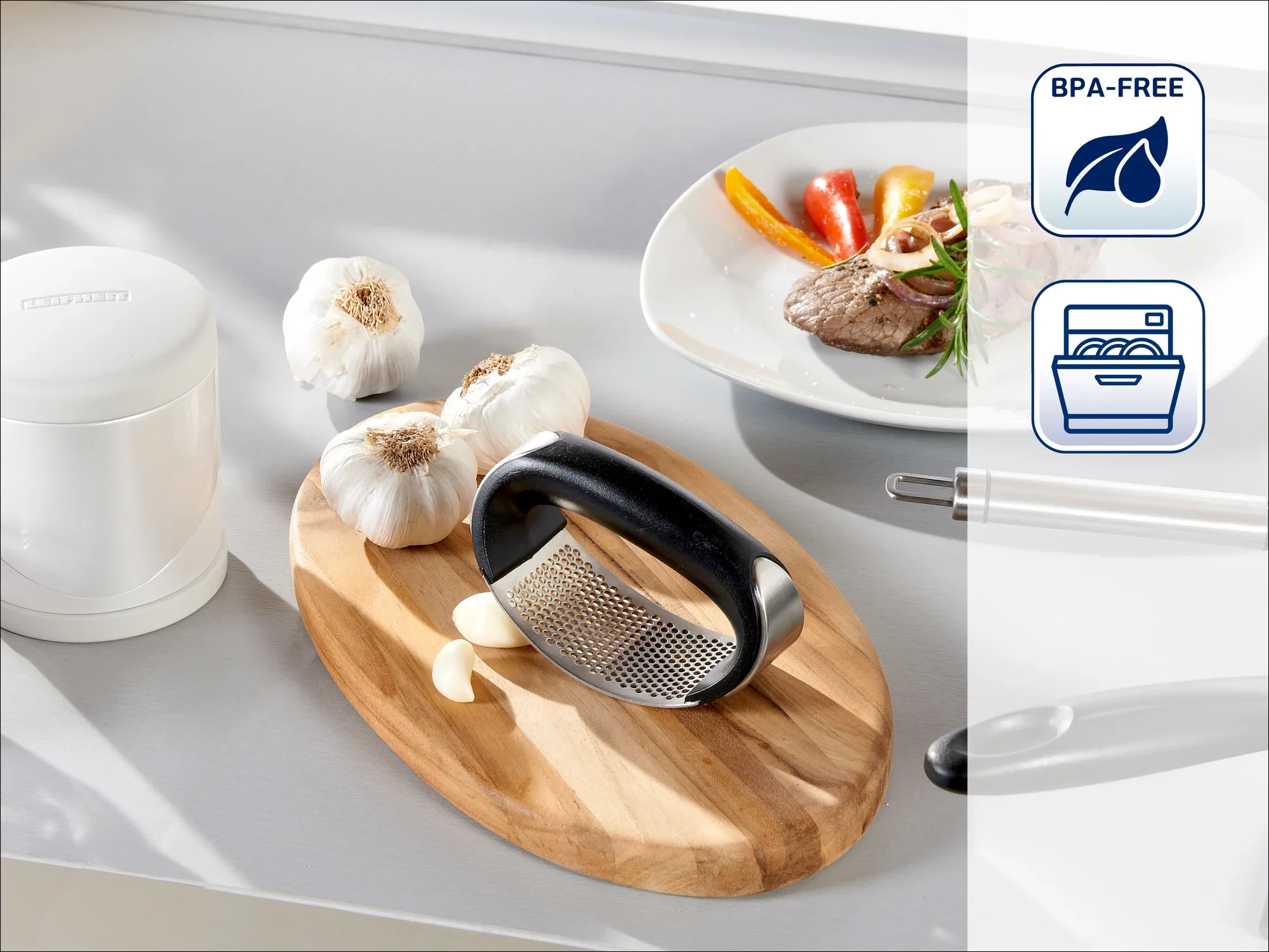 Presse à ail sur une planche en bois avec de l'ail frais. À l'arrière-plan, une assiette avec de la viande et des légumes. Les symboles indiquent que le produit est sans BPA et lavable au lave-vaisselle.