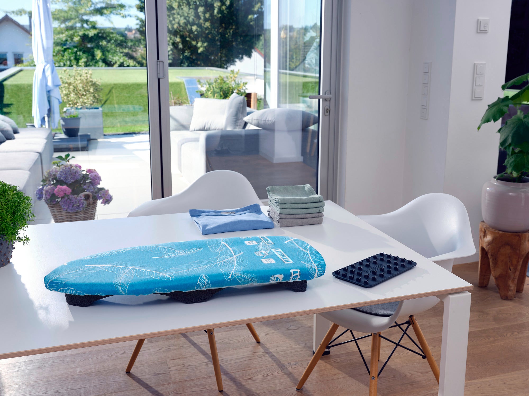 Bügeltisch Air Board Table Compact auf einem modernen weißen Tisch in einem hellen Raum mit Blick auf eine Terrasse. Der Tisch ist mit Stühlen und Pflanzen dekoriert.