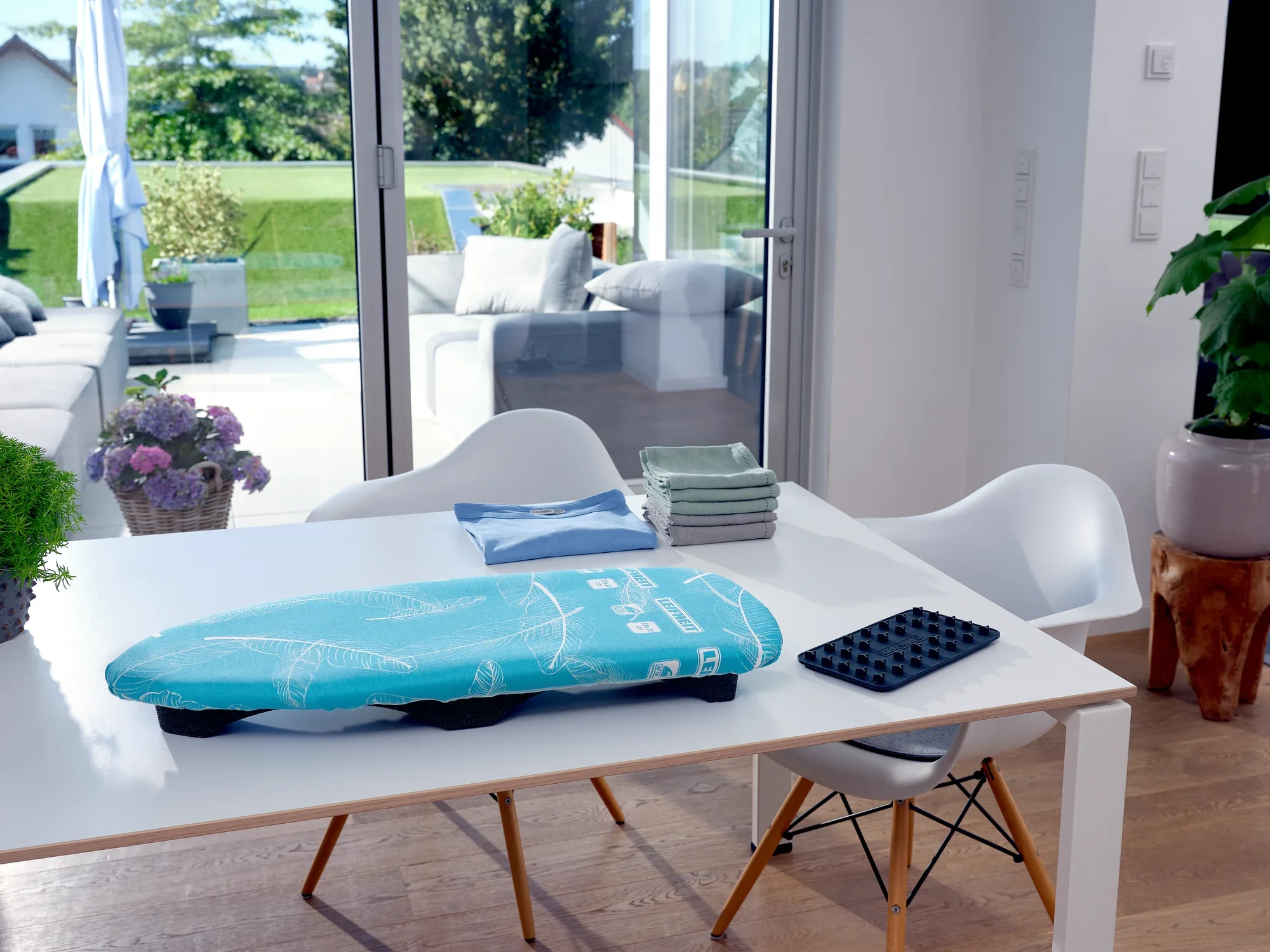 Air Board Table Compact ironing table on a modern white table in a bright room with a view of a terrace. The table is decorated with chairs and plants.