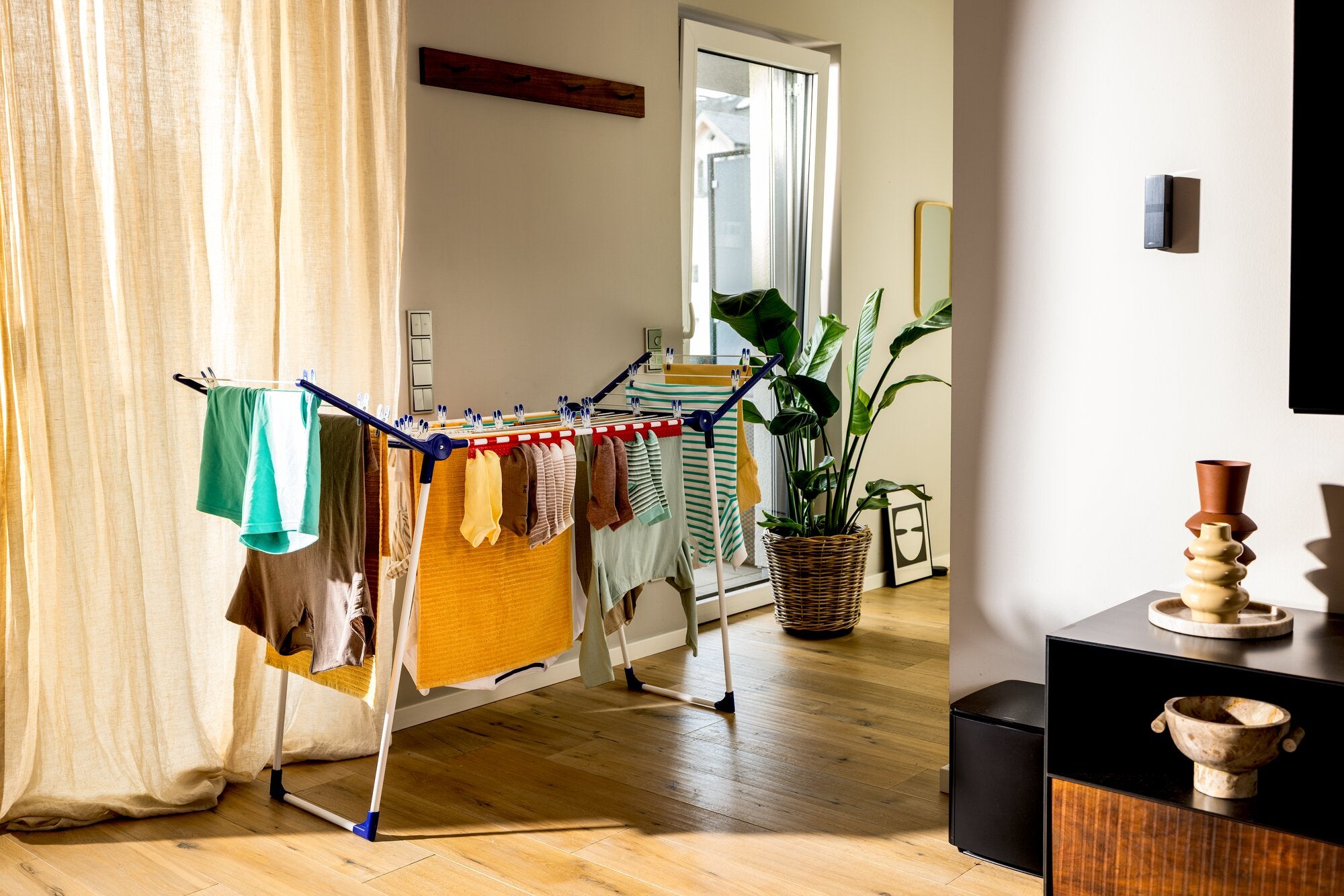 Leifheit Wäschetrockner Pegasus Slim 15 m steht mit aufgeklappten Flügeln in einem hellen Wohnraum; auf den Leinen hängen bunte Kleidungsstücke, daneben Pflanzen, Spiegel und große Vorhänge.