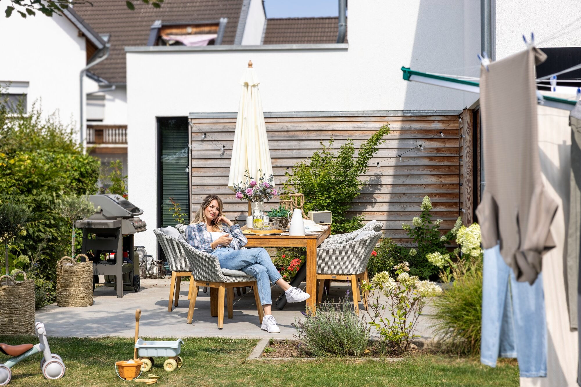 Eine Frau sitzt entspannt an einem Gartentisch, umgeben von Pflanzen und einem modernen Haus. Im Vordergrund hängt Wäsche an der Leifheit Linomatic 600 Deluxe Wäschespinne.
