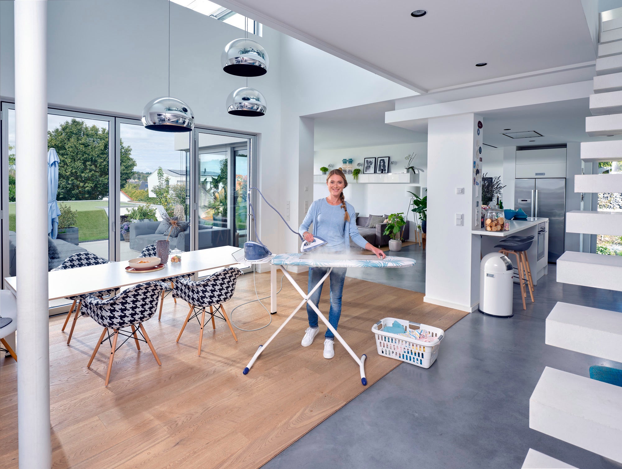 Een vrouw strijkt in een moderne, lichte woonkamer met de Air Board Express M Solid strijkplank. De kamer is stijlvol ingericht met grote ramen en een eettafel.