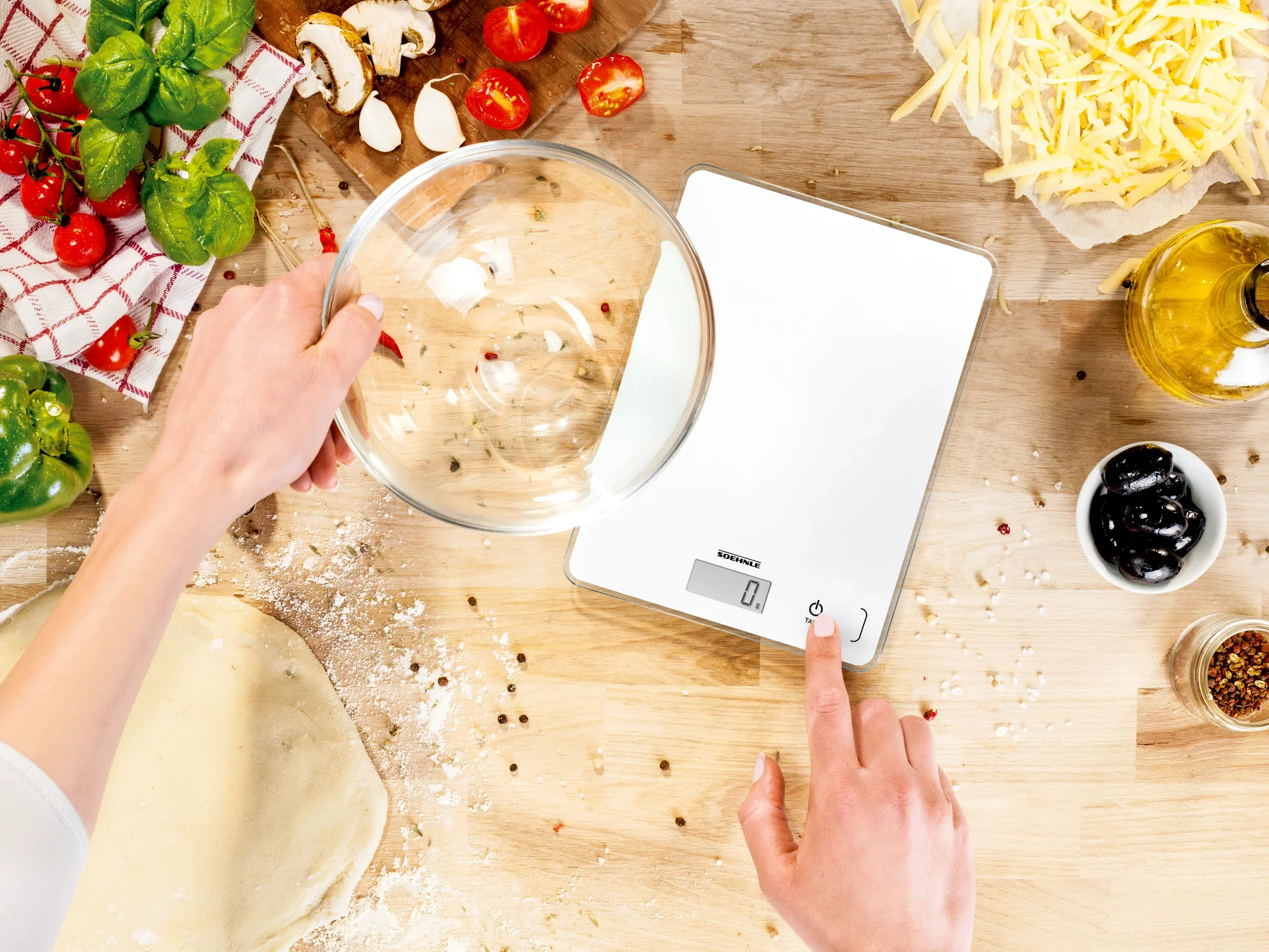 A digital kitchen scale stands on a work surface surrounded by ingredients such as tomatoes, olive oil and dough. One hand holds a bowl while the other operates the scale.