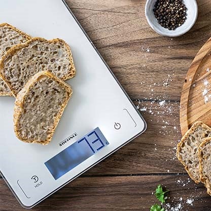 Scheiben Vollkornbrot auf einer digitalen Kuechenwaage mit 73-Gramm-Anzeige, daneben Pfeffer in einer Schale und Holzbrett mit Brot.