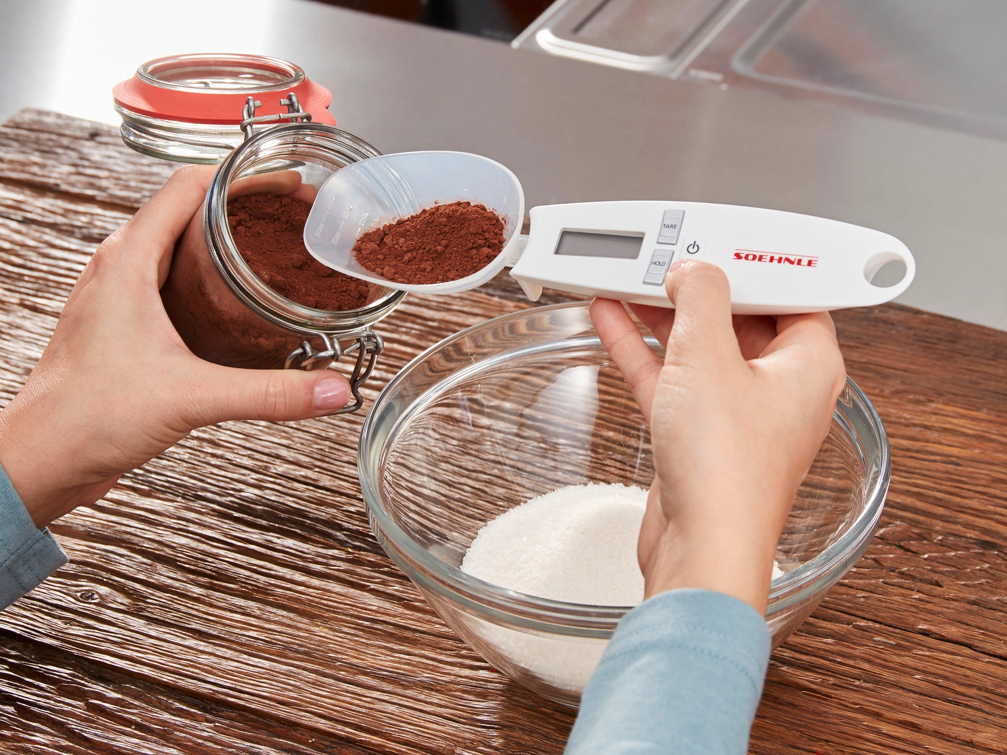 Eine Person verwendet die digitale Küchenwaage Cooking Star, um Kakaopulver aus einem Glas in eine Schüssel mit Zucker zu wiegen. Die Waage zeigt präzise Messwerte an.