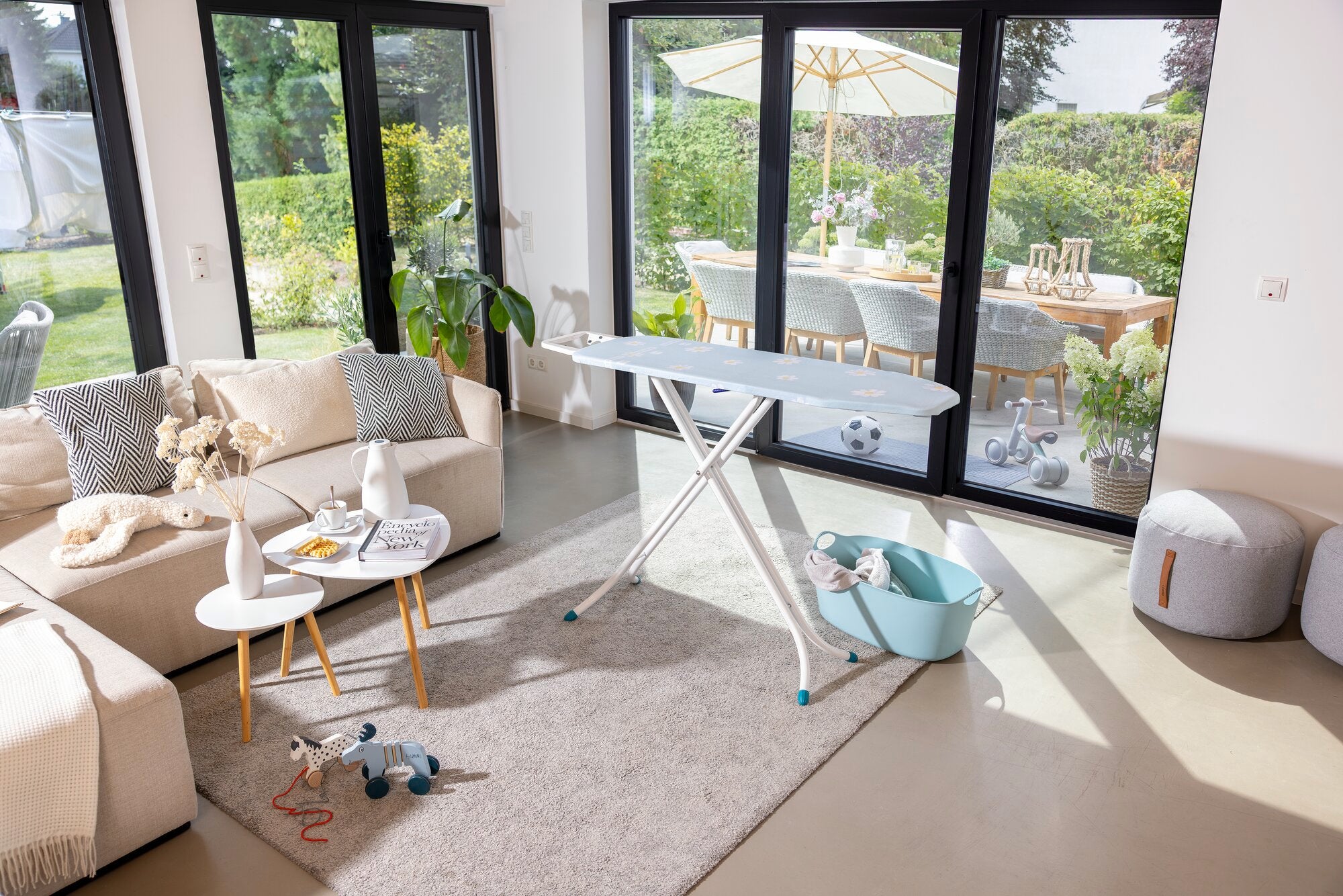 Bügelbrett Leifheit Classic M Compact steht in einem hellen Wohnzimmer mit großen Fenstern. Es ist umgeben von modernen Möbeln und Pflanzen, die eine gemütliche Atmosphäre schaffen.