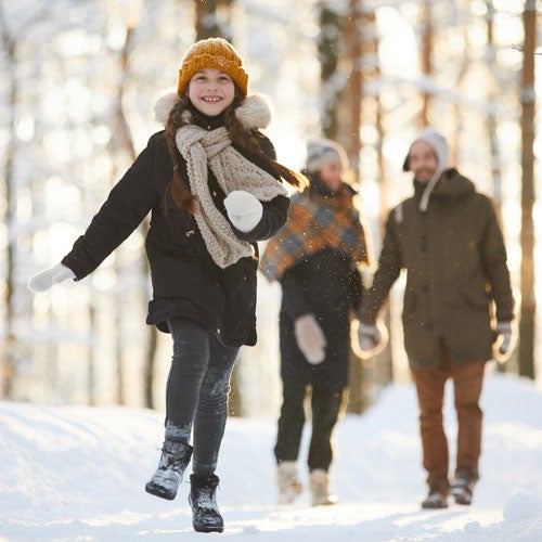 Ein lachendes Maedchen mit gelber Muetze und Schal laeuft im Schnee; zwei Erwachsene folgen im Hintergrund durch einen verschneiten Wald.