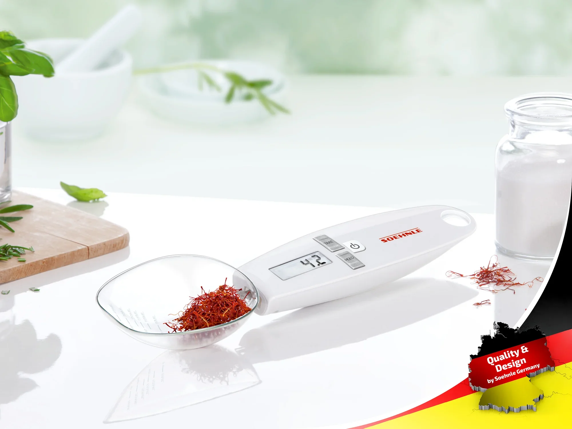 Cyfrowa łyżka miarowa firmy Soehnle wskazuje 4,3 grama szafranu. Waga jest biała z wyraźnym wyświetlaczem i jest umieszczona na blacie kuchennym. W tle widoczne są zioła i szklanka.