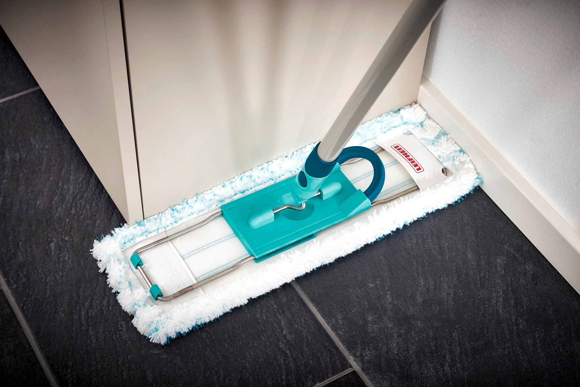 Un balai Profi XL avec un manche en acier est utilisé dans un coin d'une pièce. Le balai est équipé d'un tampon de nettoyage vert et est idéal pour un nettoyage en profondeur.