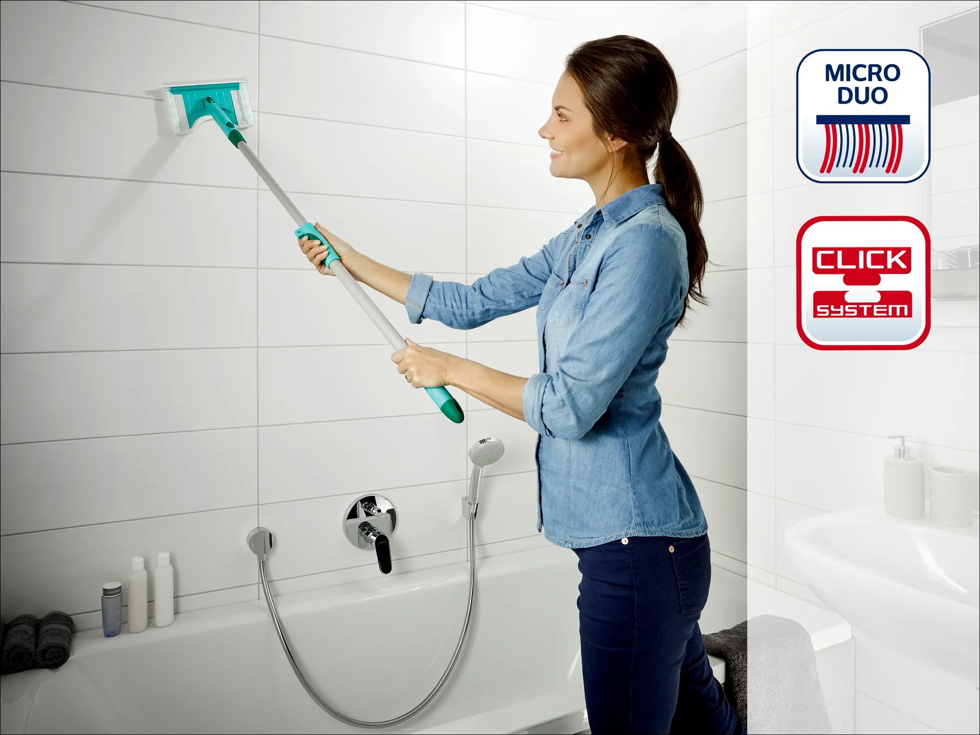 Kobieta czyści ścianę wyłożoną kafelkami w łazience za pomocą systemu Leifheit Click i ściągaczki Micro Duo. Ściągaczka ma długi uchwyt zapewniający wygodną pracę.