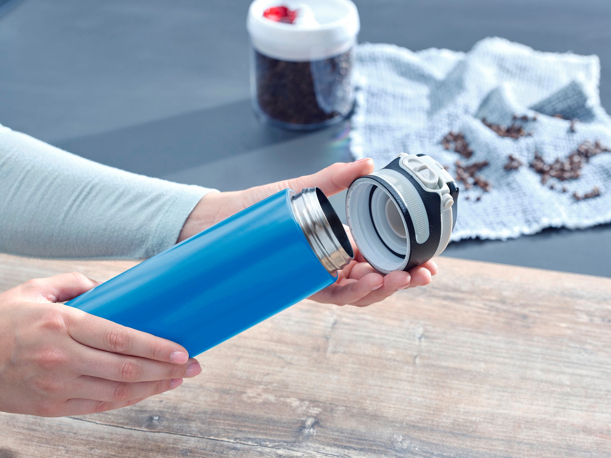 Isolierbecher Flip 600ml in Hellblau wird geöffnet. Der Becher liegt auf einem Holztisch, im Hintergrund sind Kaffeebohnen und ein Behälter zu sehen. Ideal für unterwegs.