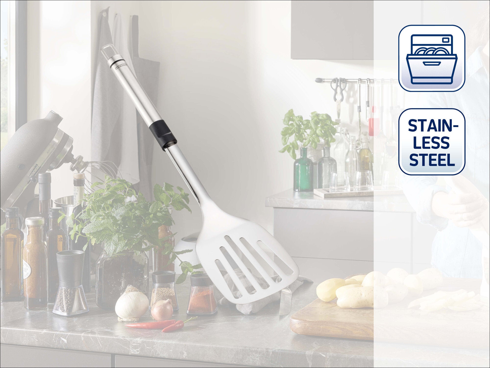 Leifheit Küchenhelfer Sterling Wender aus Edelstahl mit breiter, geschlitzter Fläche und ergonomischem Griff, dekorativ an eine Küchenwand gelehnt in heller, moderner Kochumgebung.