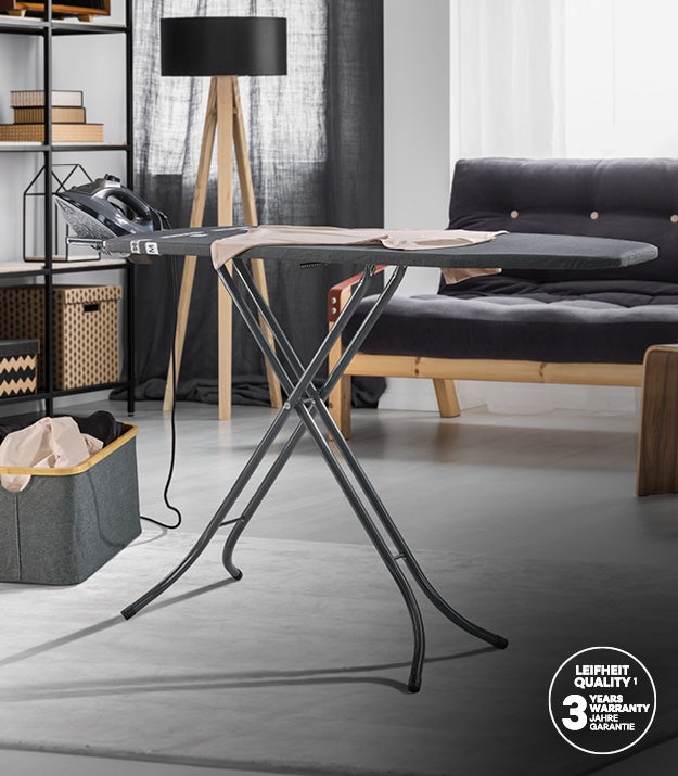 Ein Buegelbrett mit Buegeleisen steht in einem modern eingerichteten Wohnzimmer mit Regal, Stehlampe und grauem Sofa.