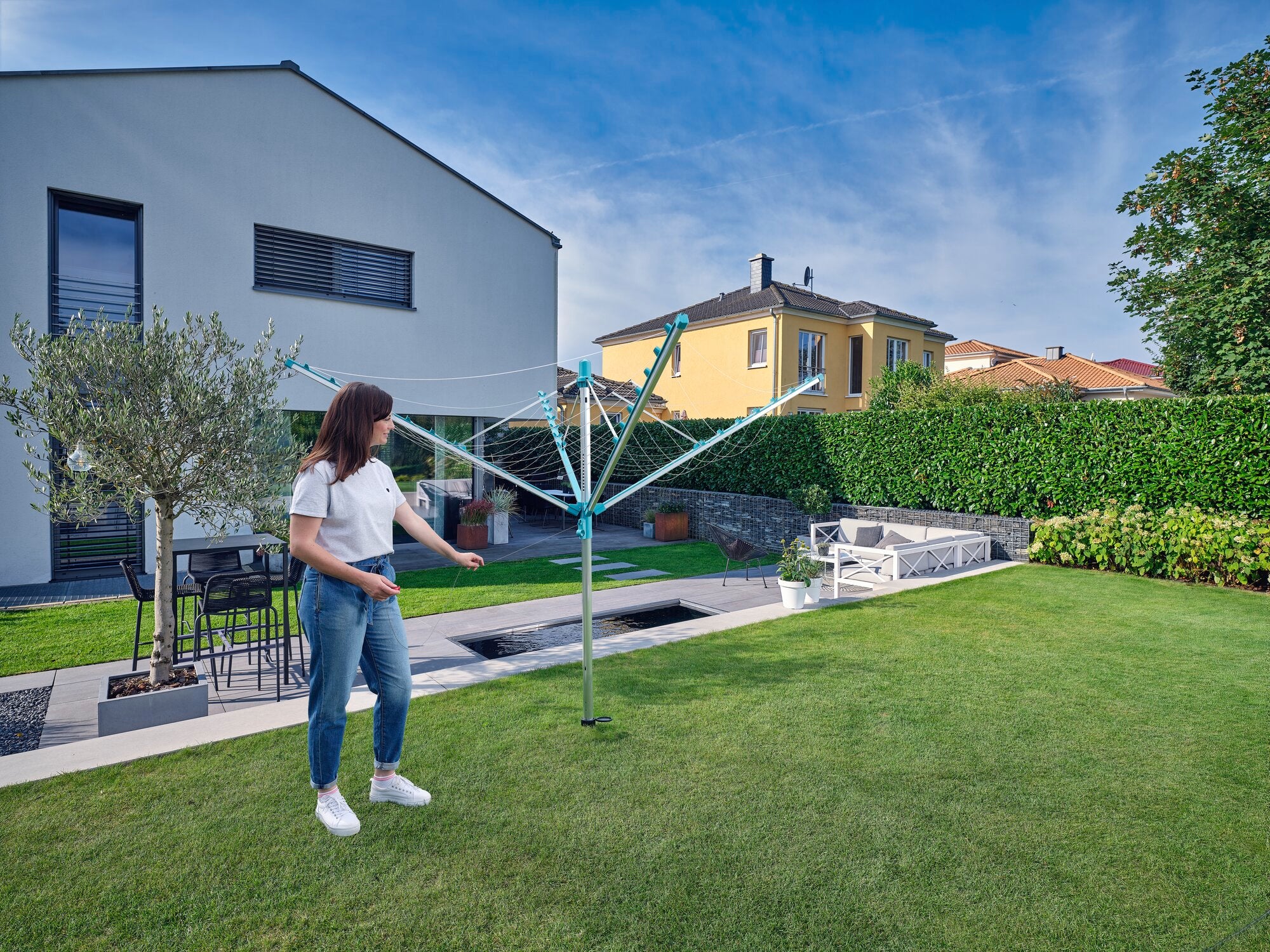 A woman stands in a well-kept garden using the Linomatic 400 Easy clothes horse. Modern houses and a hedge can be seen in the background. The sky is clear and blue.