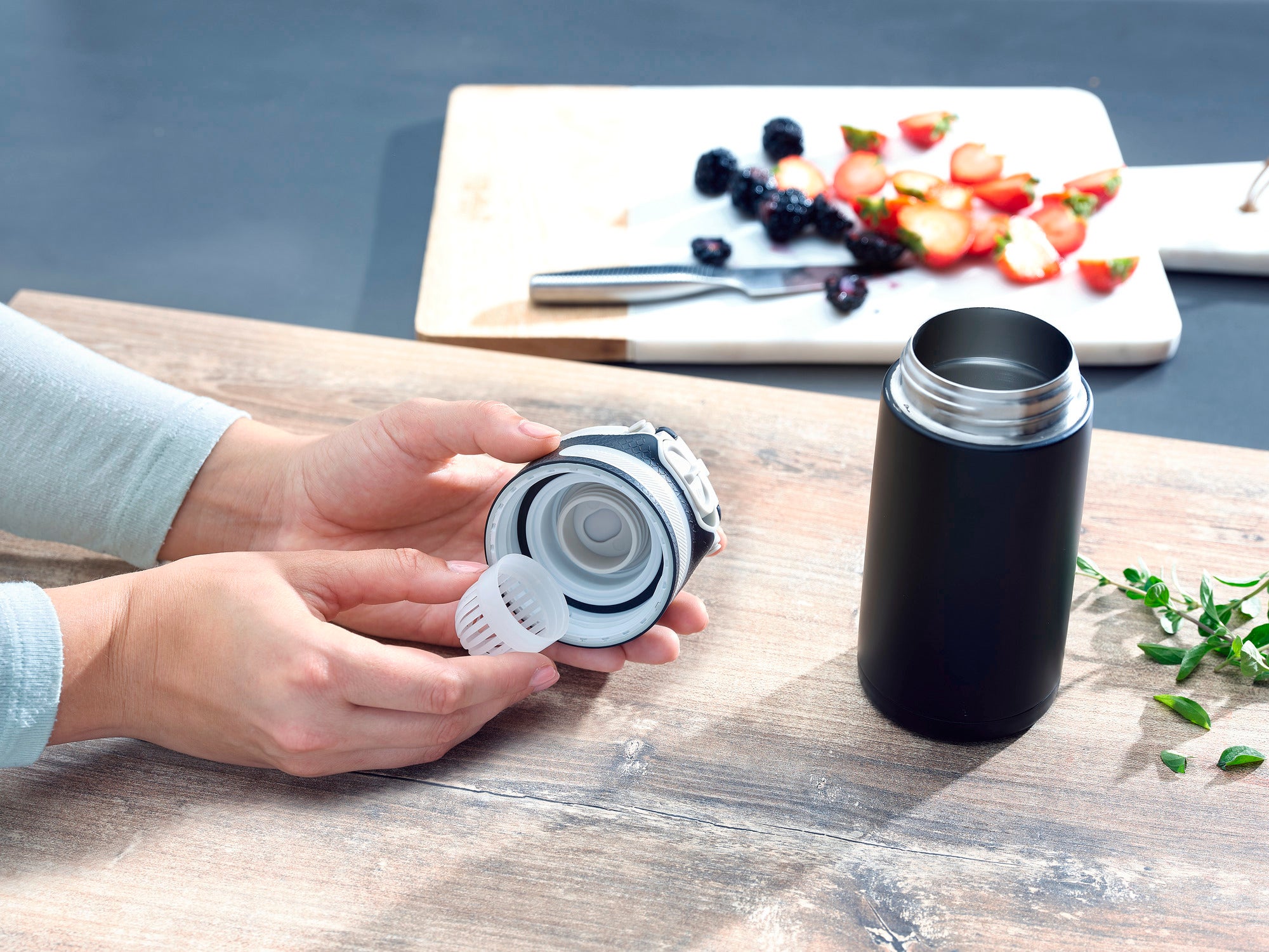 Flip 350 ml insulated mug in black on a table. A person is holding the lid of the mug, while a cutting board with fruit can be seen in the background. Ideal for on the go.