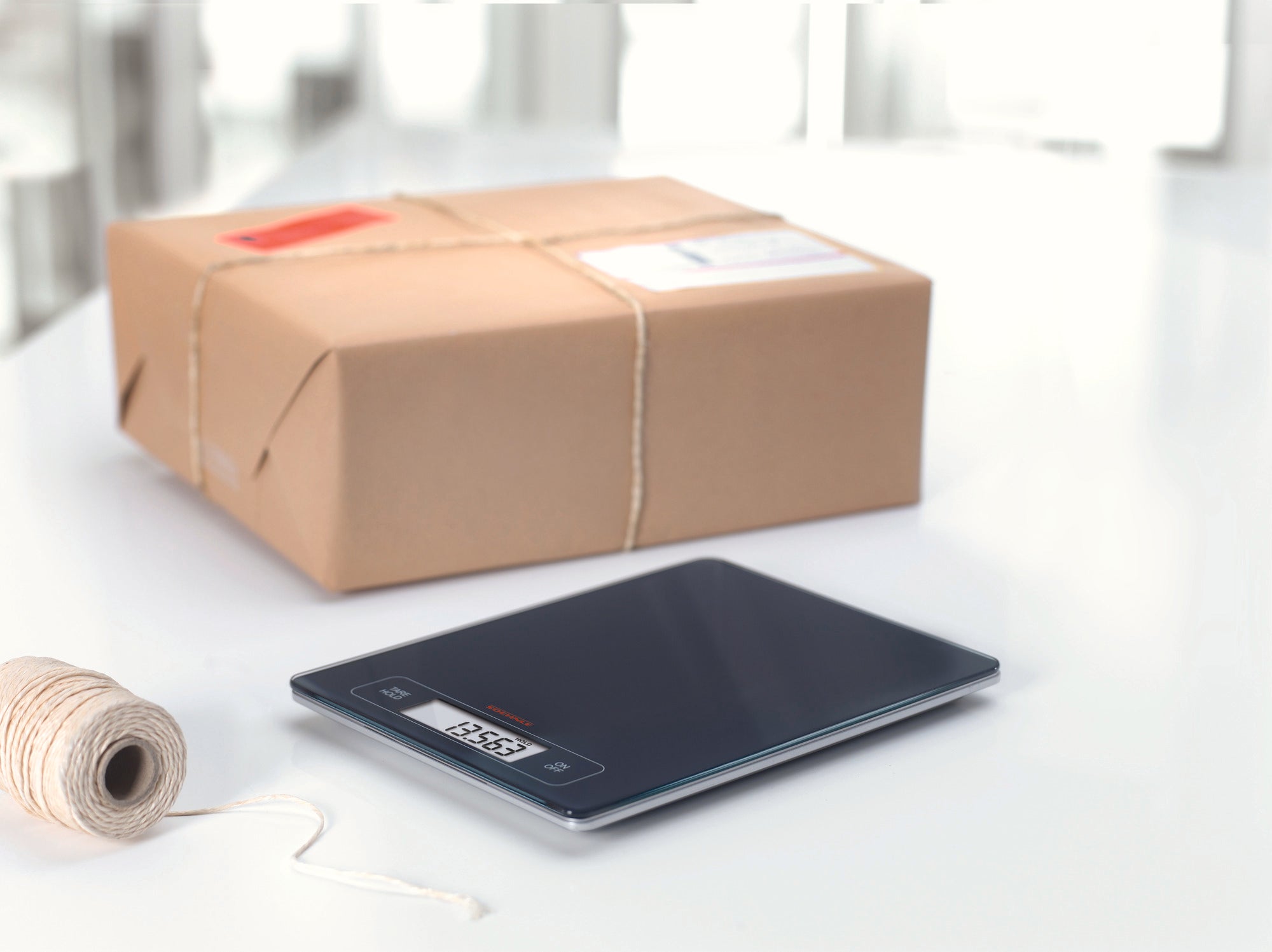 Eine digitale Küchenwaage liegt auf einem weißen Tisch neben einem Paket und einer Rolle Schnur. Die Waage zeigt ein Gewicht an und hat ein modernes, flaches Design.