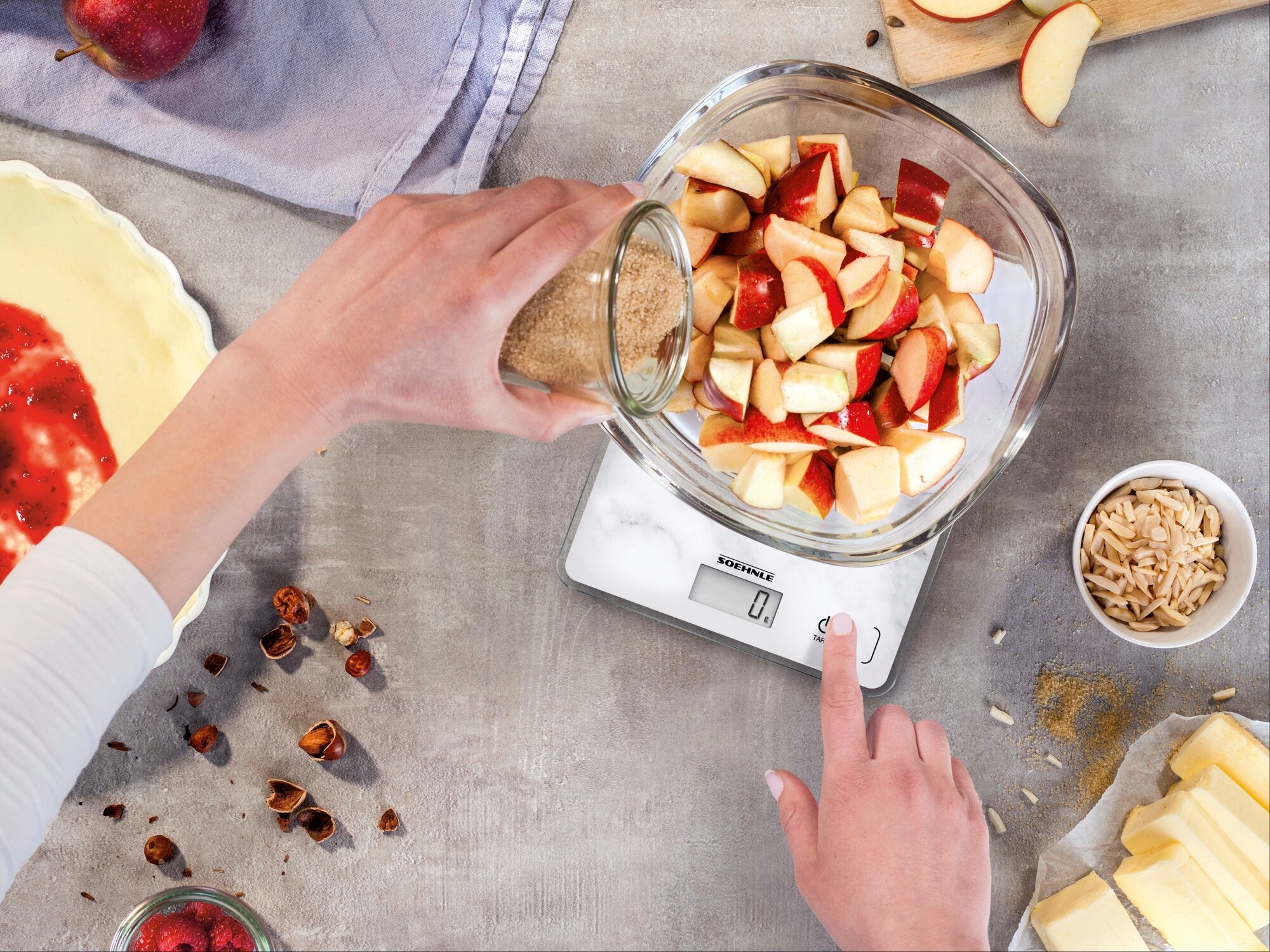 Eine Person wiegt Apfelstücke in einer Glasschüssel auf der digitalen Küchenwaage Page Compact 300 Marble Tara. Die Waage zeigt das Gewicht präzise an, während Zutaten für ein Rezept vorbereitet werden.