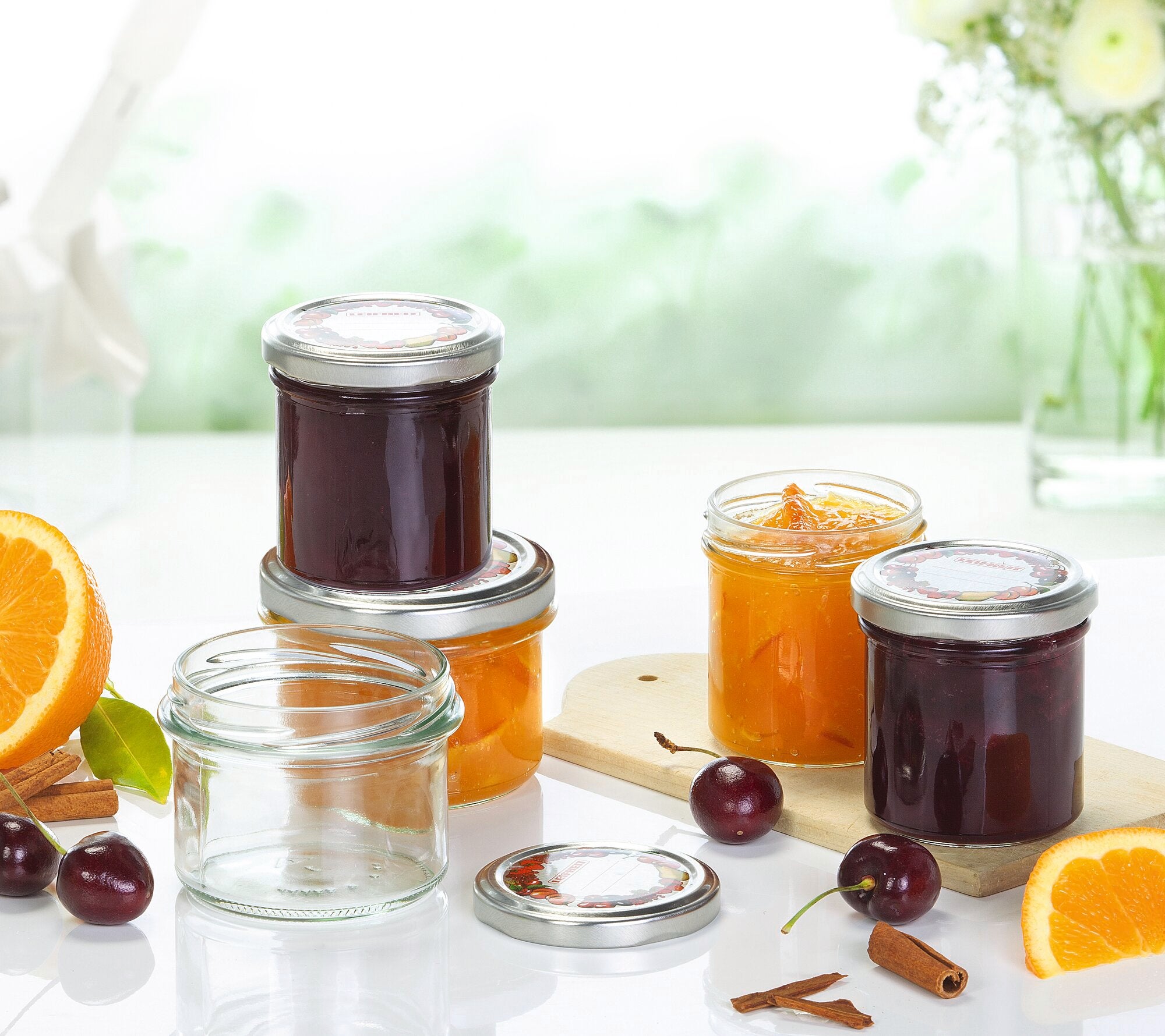 Sturzglaeser mit Marmelade und leeren Gläsern auf einem Tisch. Im Hintergrund sind Blumen und eine verschwommene grüne Landschaft zu sehen. Frische Orangen und Kirschen liegen daneben.