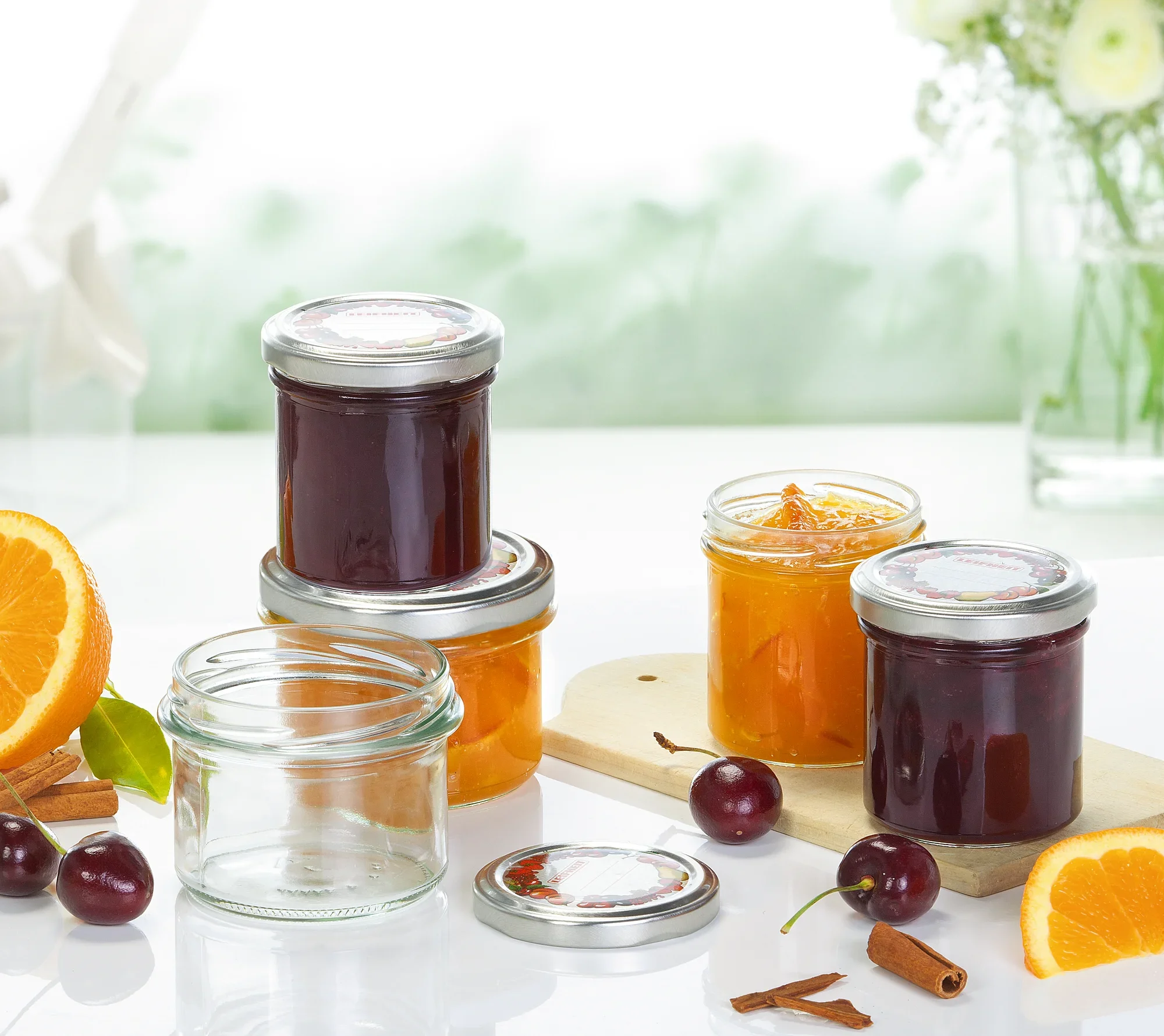Tumblery z dżemem i puste słoiki na stole. W tle widać kwiaty i rozmyty zielony krajobraz. Obok leżą świeże pomarańcze i wiśnie.